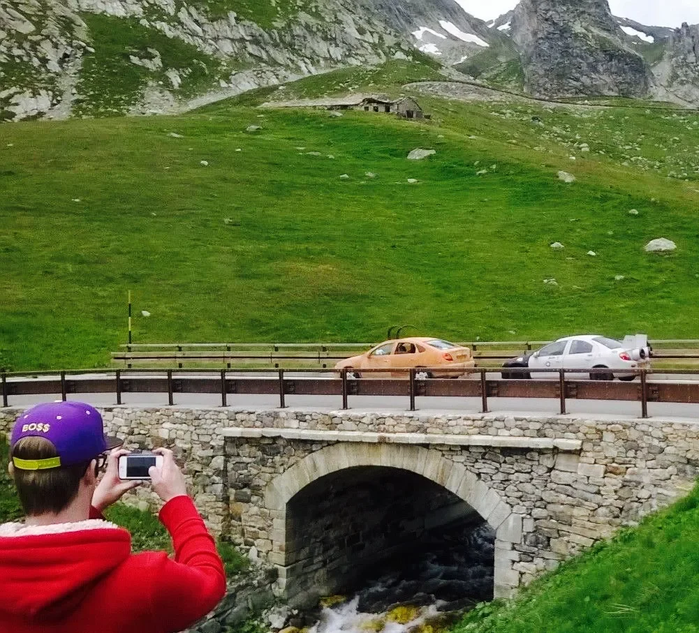 A person in a red hoodie and purple cap taking a photo of a stone bridge over a creek. In the background, there is a green hillside with two cars, one orange and one white, driving across a road on the hill. Farther back, there are rocky mountains.