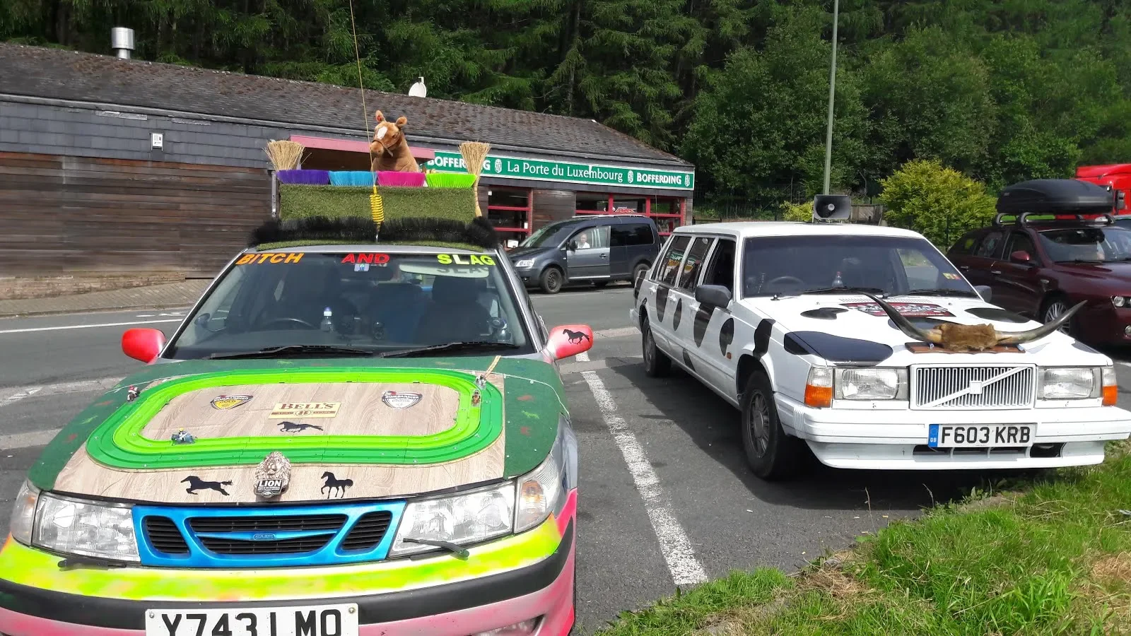 Parking lot with cars decorated for a feline-themed event; one car has a lion or cheetah figure on the hood and animal decals, another has a horned animal figure on the front, and a float with a lion's head and a laurel wreath is visible in the backg
