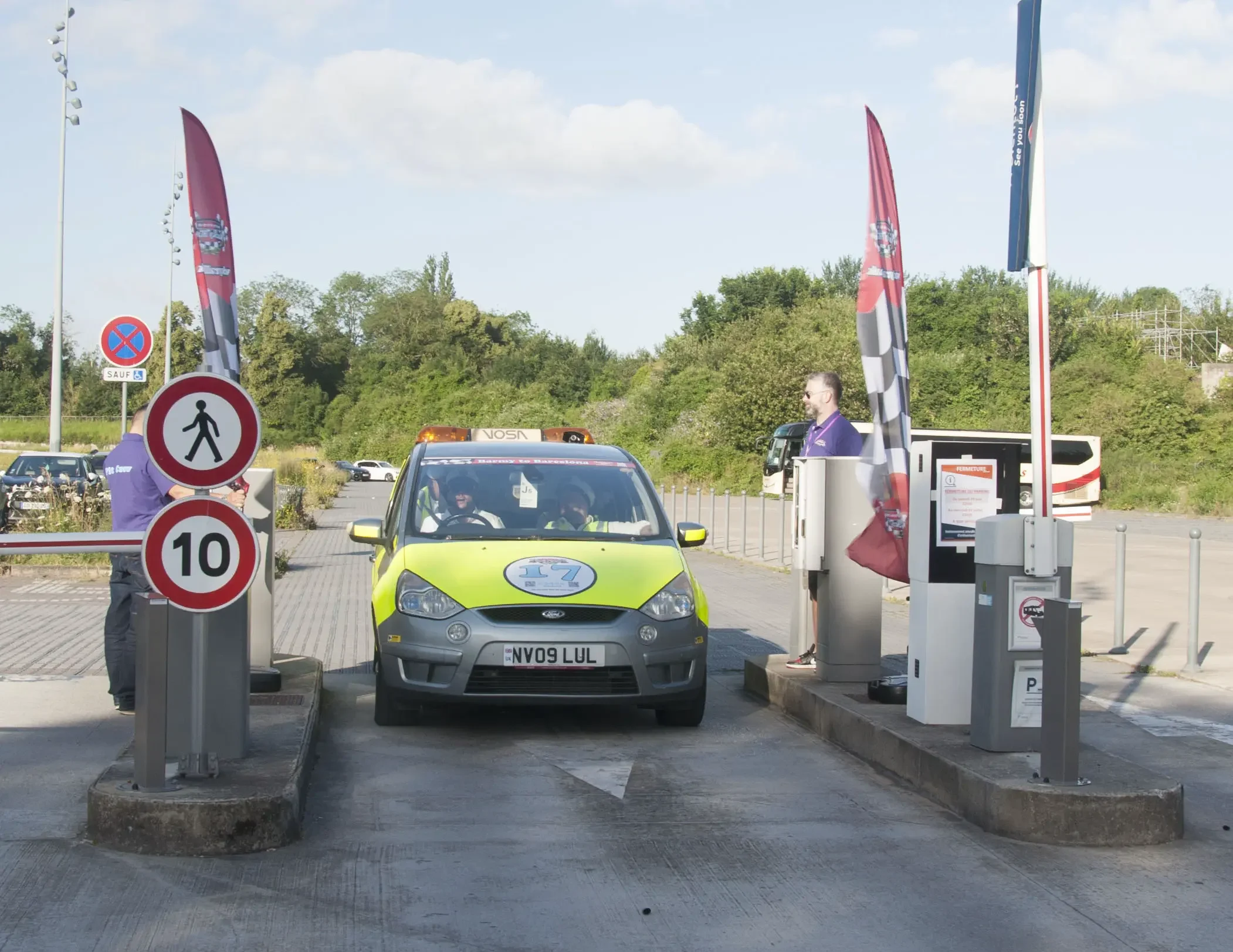 A green and gray driving school car with the license plate NV09 LUL passing through a parking gate, driven by a person wearing a black cap and sunglasses, on a sunny day with a blue sky and green trees in the background.