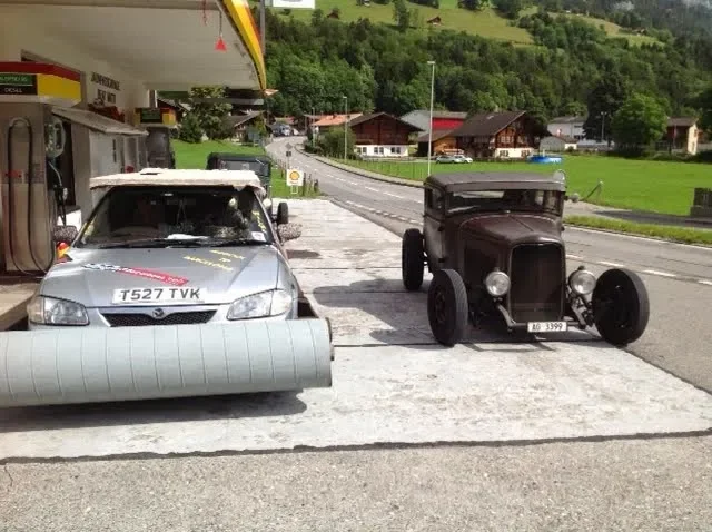 A vintage black car parked next to a modern silver car with what appears to be a large pipe or vent cover on the front, at a gas station in a rural area with green hills, trees, and houses in the background.