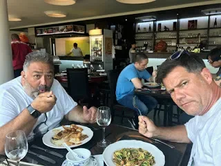 Two men dining at a restaurant with plates of food and drinks on the table, with other diners and a bar in the background.