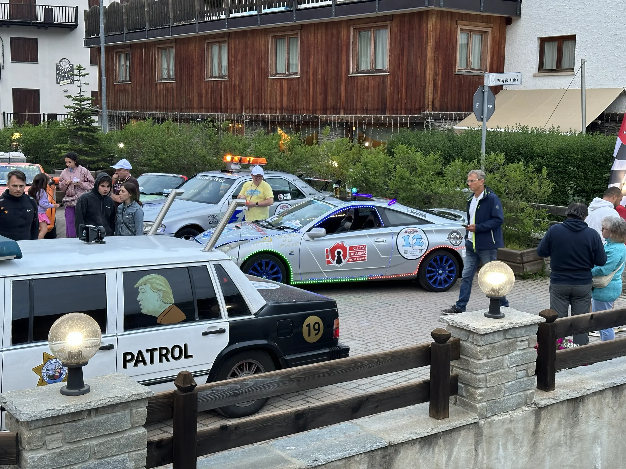 A gathering of people near a police patrol car and a decorated sports car parked outside a building. The patrol car is white with a caricature of Donald Trump on the side and the word 'PATROL'. The sports car is gray with blue wheels and colorful LED