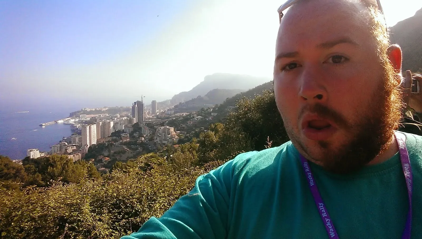 A man taking a selfie on a hillside overlooking a coastal city with tall buildings and mountains in the background.