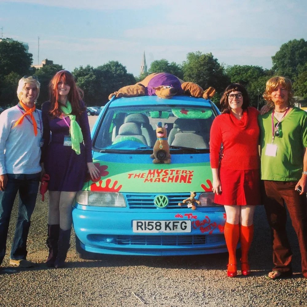 Group of five people standing in front of a colorful Volkswagen car decorated as "The Mystery Machine" from Scooby-Doo, with Scooby-Doo plush toys on the hood and a person dressed as a cartoon character lying on the roof.