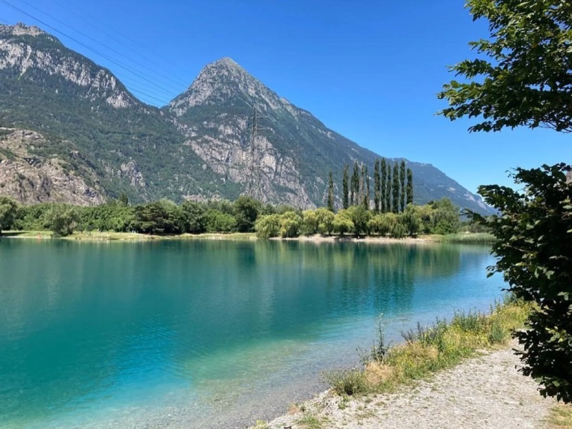 A peaceful lake with clear blue water reflecting green trees along the shore and towering mountain cliffs in the background under a bright blue sky.