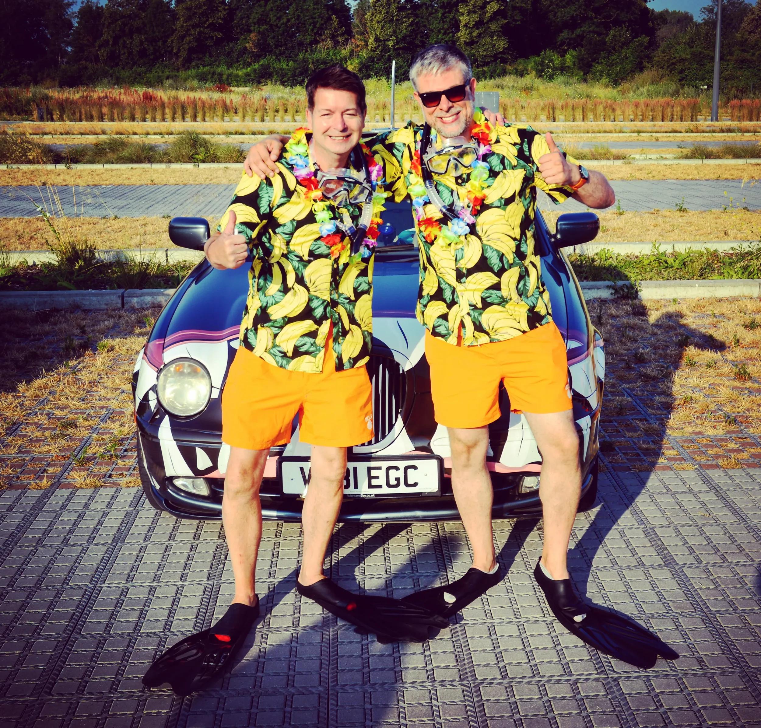 Two men in matching tropical shirts, orange shorts, and snorkeling gear standing in front of a decorated car, giving thumbs up, on a sunny day.
