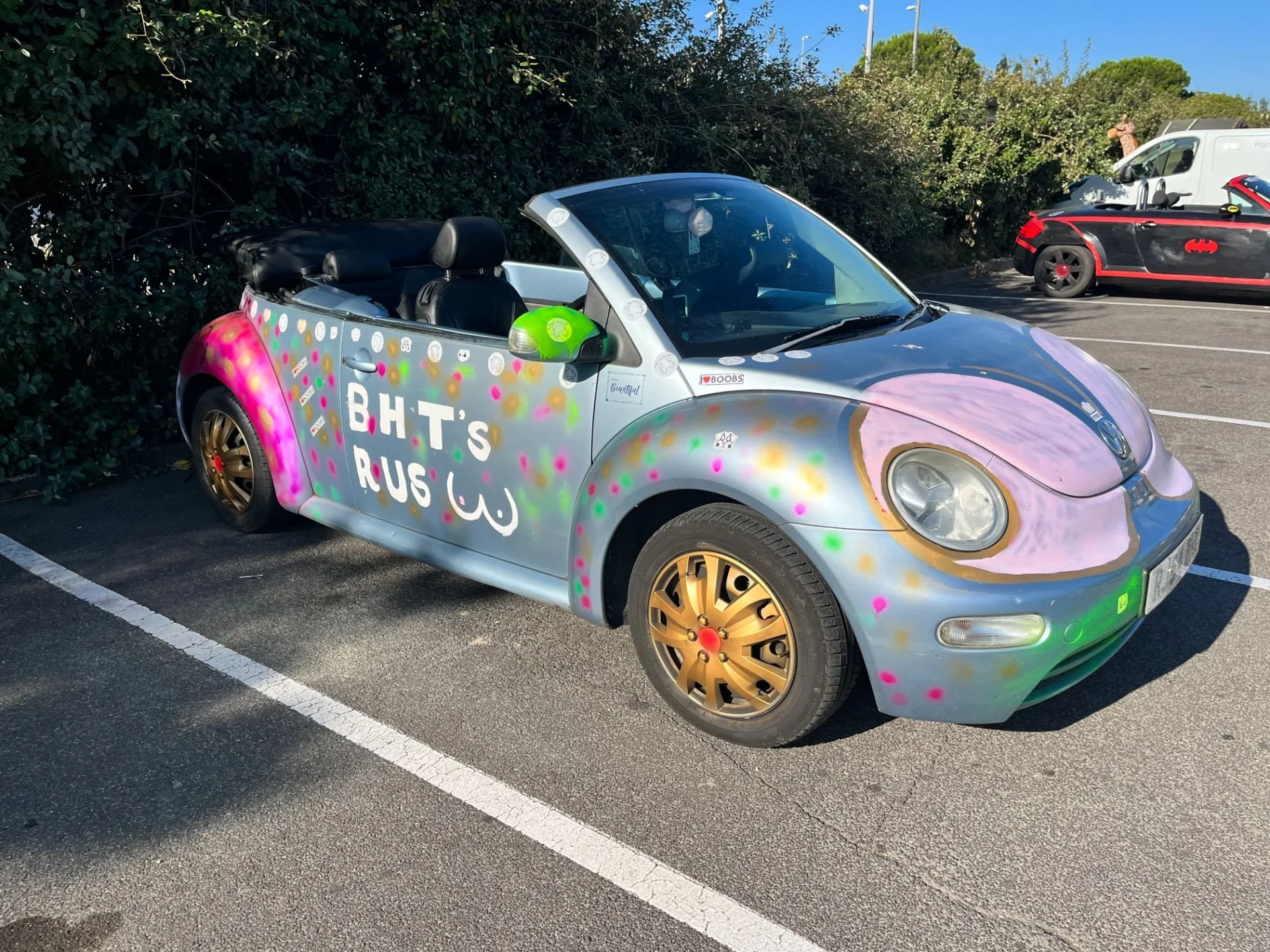 A colorful, graffiti-style painted convertible car with gold wheels, parked in a parking lot beside a hedge. The car has the words 'BHT'S RUSH' written on its side and features pink, purple, green, and gold spray paint accents.