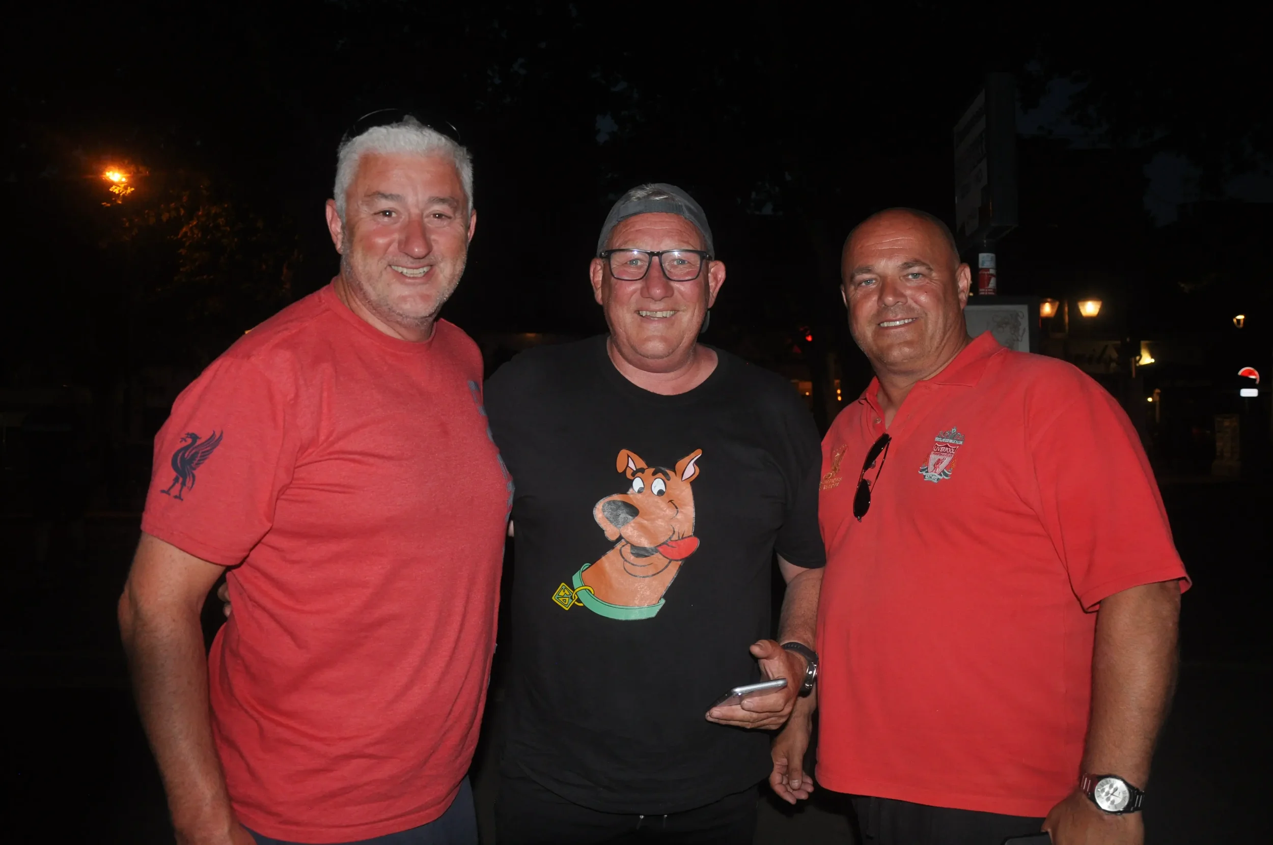 Three middle-aged men standing close together outdoors at night, smiling for the camera, wearing casual t-shirts, two in red and one in black featuring Scooby-Doo.