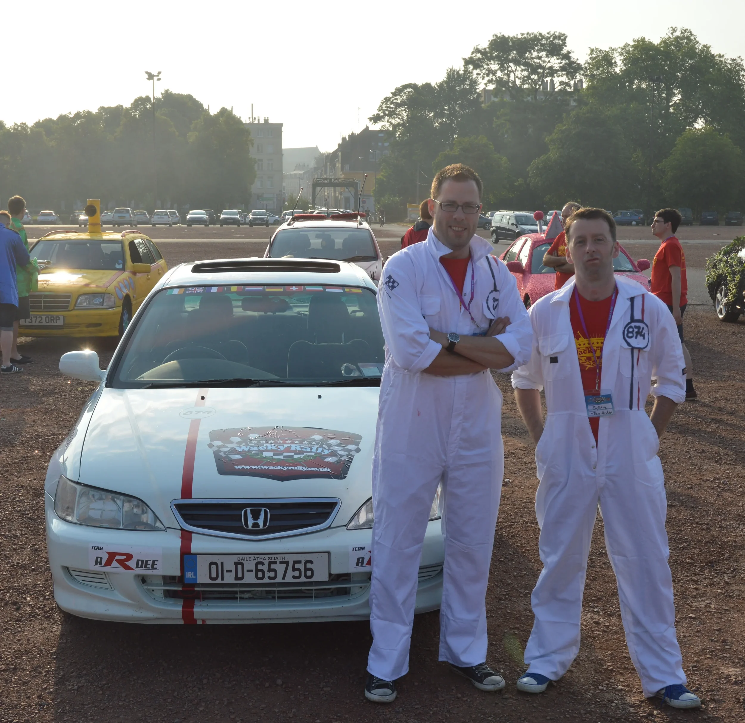 Two men in white racing suits standing next to a decorated Honda car at a rally event, with more cars and people in the background.