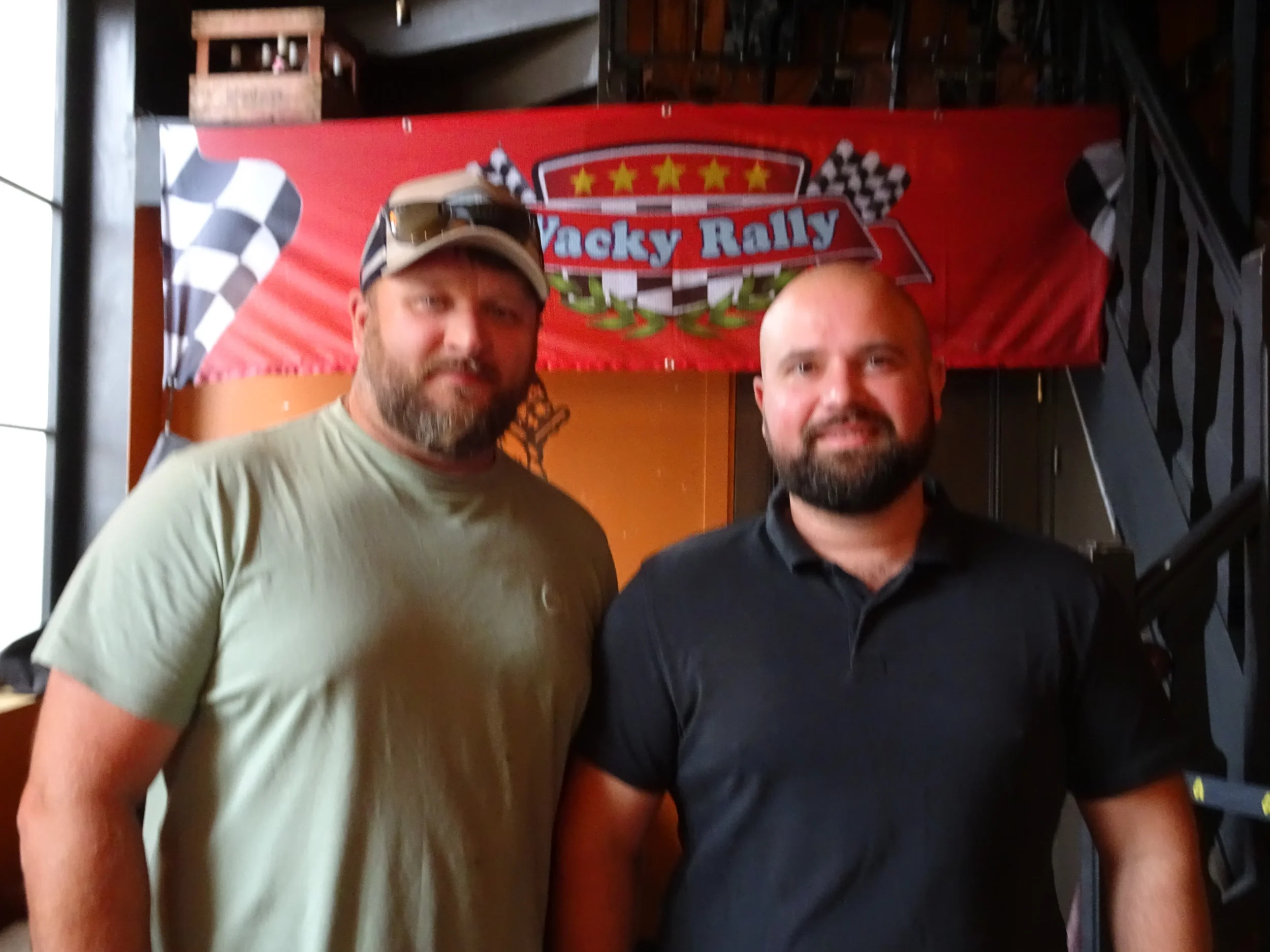 Two men standing side by side indoors, in front of a red banner with checkered flags and the words 'Trucky Rally' on it. The man on the left has a beard, wears a tan cap with sunglasses resting on it, and a light olive green t-shirt. The man on the r