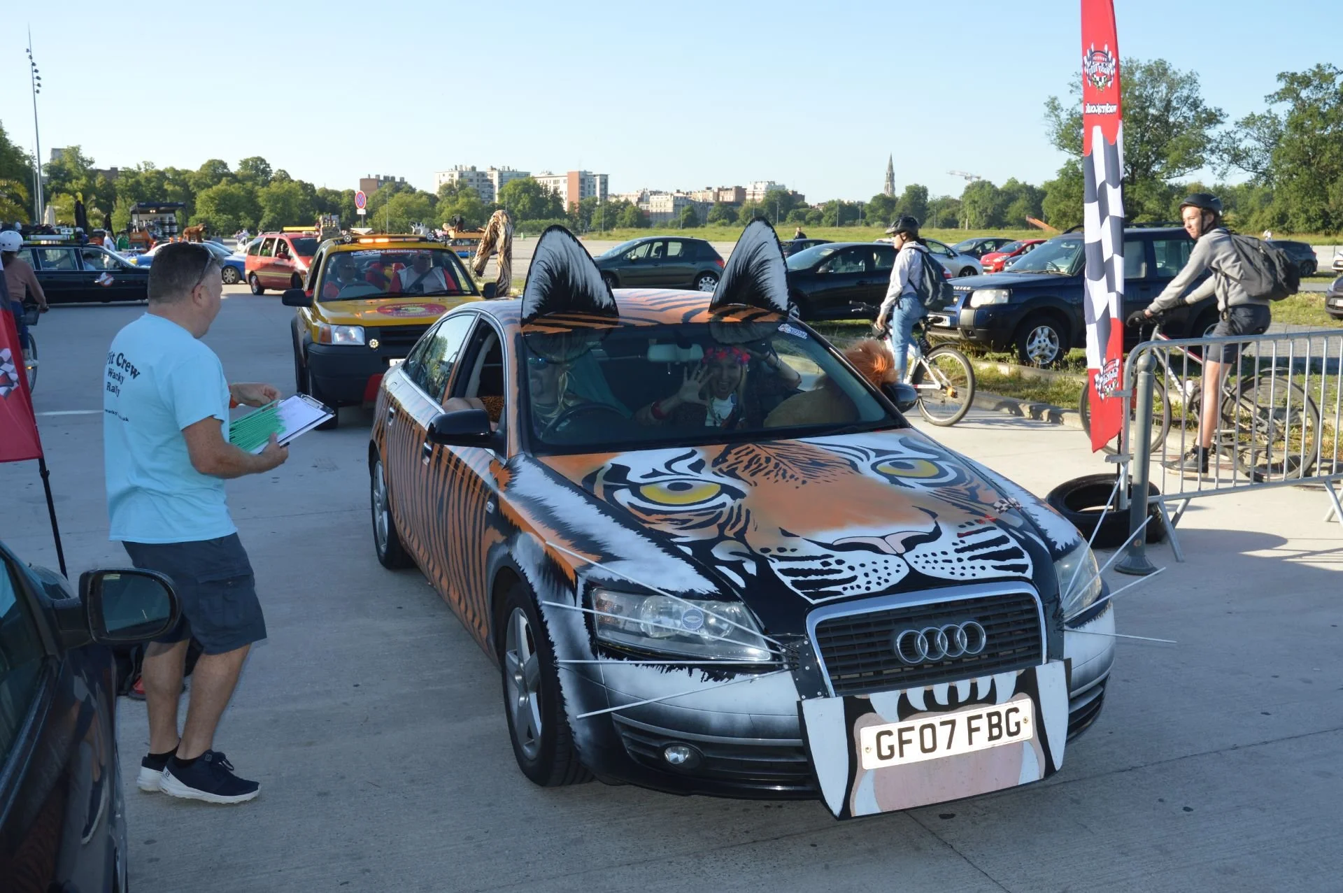 A car decorated with a tiger face design and large black ears on the roof, displayed at an outdoor event with people, bicycles, and cars around.