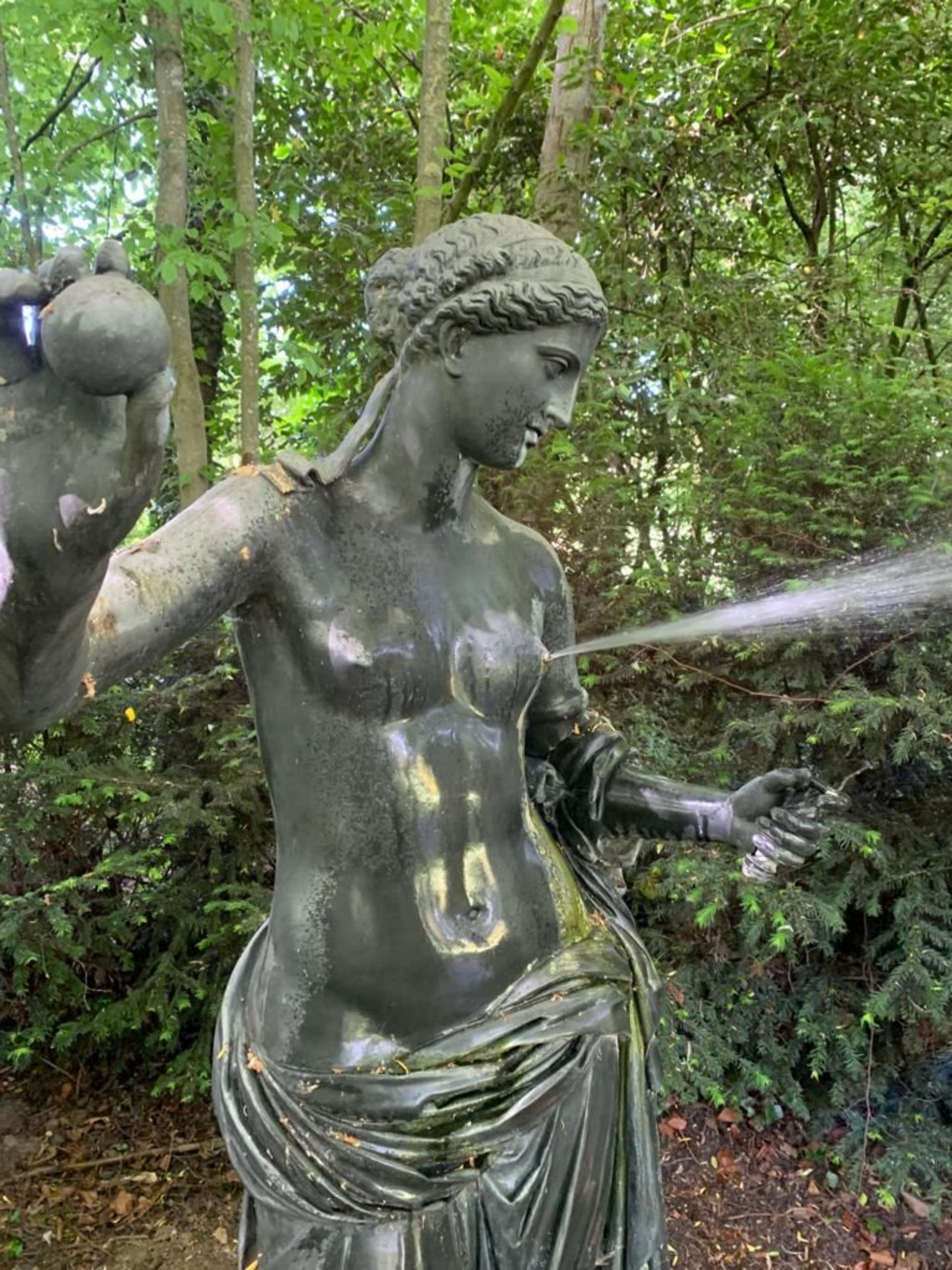 A metal statue of a woman holding a watering can, watering a plant, surrounded by trees and greenery.
