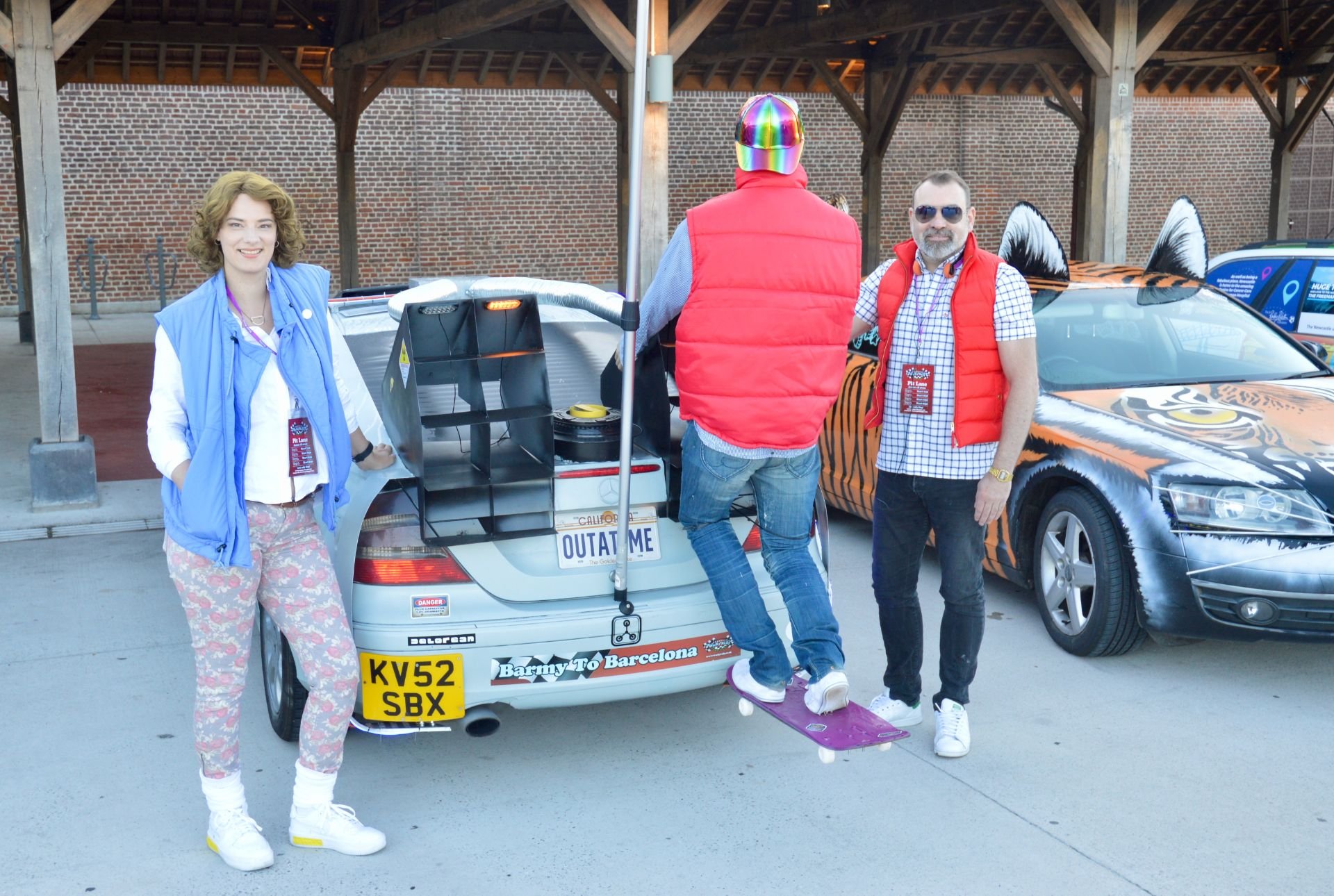 Three people stand beside a small vehicle decorated with a 'Back to the Future' theme. One woman is smiling, wearing a blue vest and patterned pants. A man with sunglasses and a red puffy vest stands on the right. Another man, wearing a red jacket wi