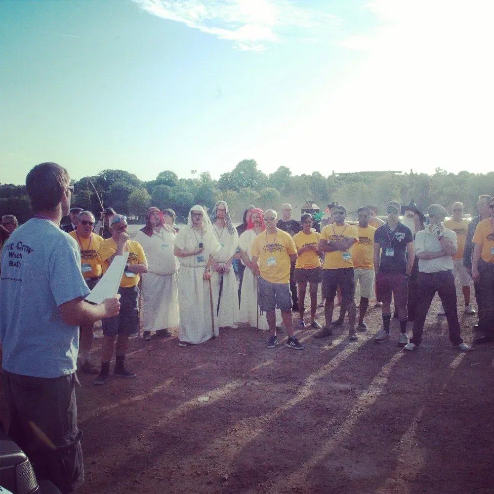 Group of people gathered outdoors on dirt ground for a speech or presentation, with some dressed in traditional Middle Eastern attire and others in yellow and black T-shirts, under a partly cloudy sky.