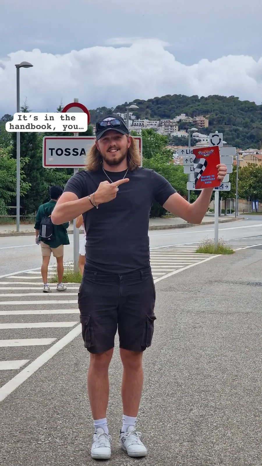 A man with long hair and a beard wearing a black t-shirt, black shorts, white sneakers, and sunglasses, standing on a street holding a booklet for the 2024 World Rally Championship in Barcelona. He is smiling and pointing to the booklet with his righ