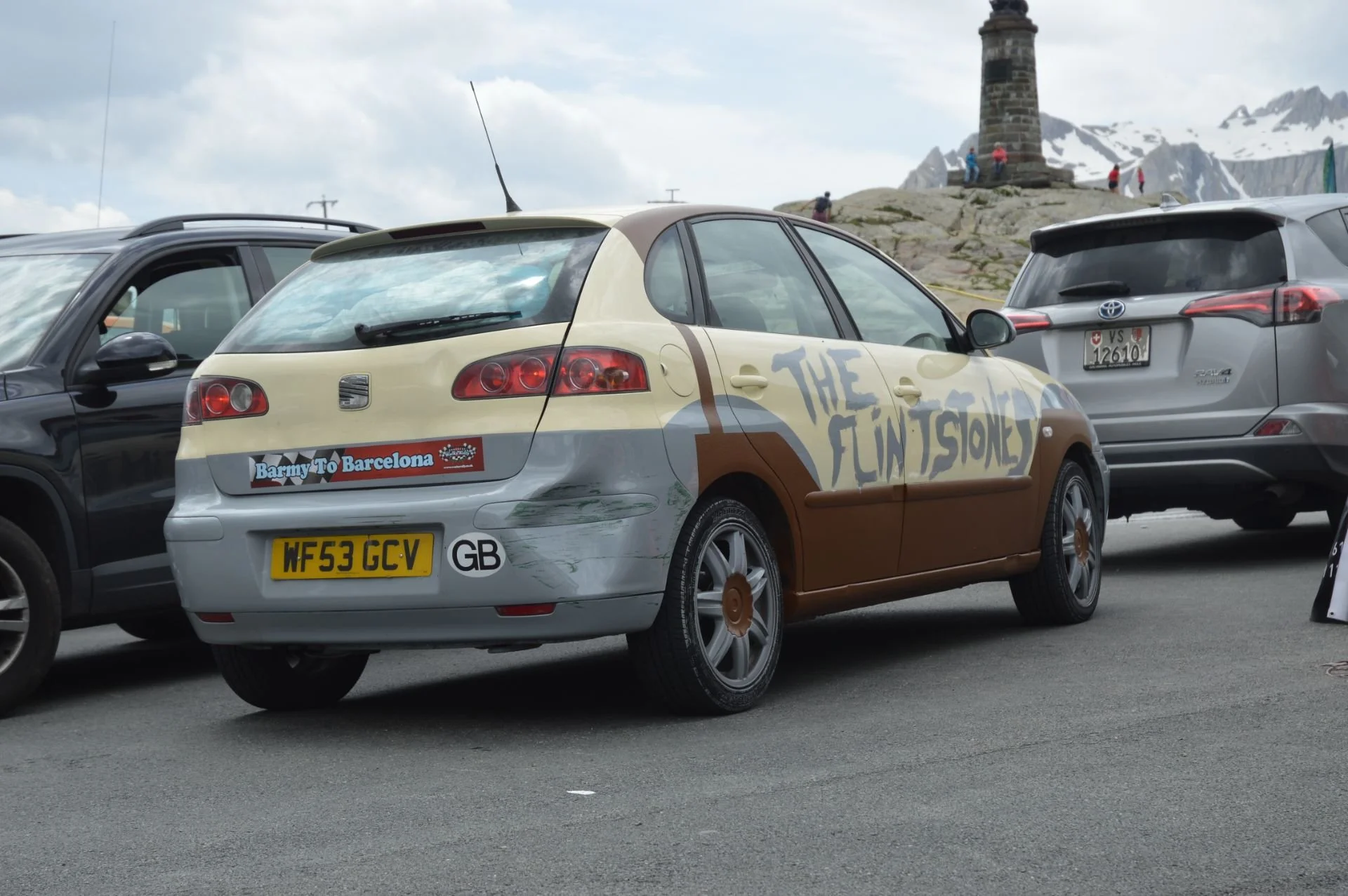 A station wagon decorated with scenes and text for 'The Flintstones,' featuring a brown and beige color scheme, parked among other cars at a scenic mountain location with a monument and snow-capped peaks in the background.