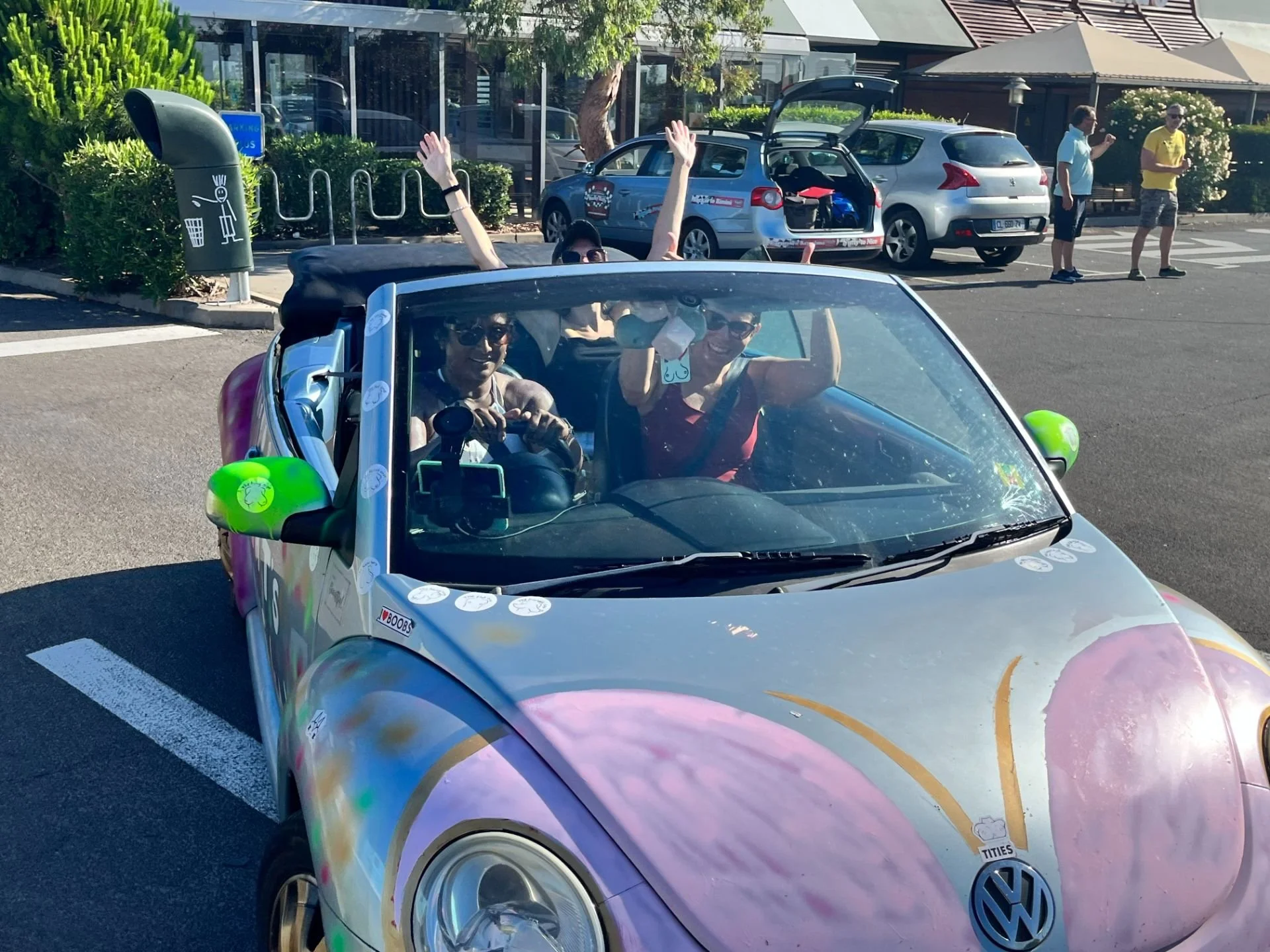 A colorful Volkswagen convertible with a rainbow design, parked in a lot. Three people are inside the car, with two women in front and one person in the back, all smiling and waving. The background shows a parking lot with other cars, a few pedestria