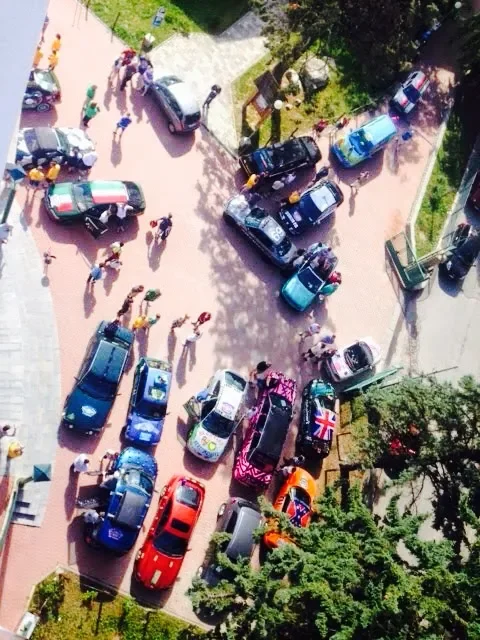 An aerial view of a street with numerous parked cars and a crowd of people walking on the sidewalk and street.