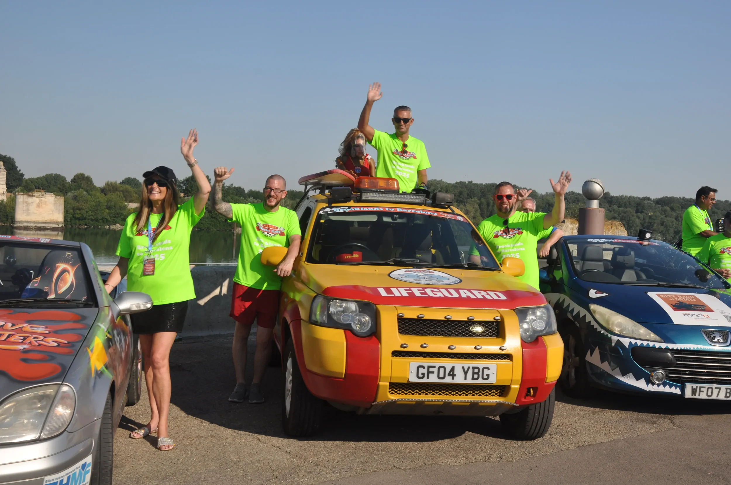Group of people in bright green T-shirts standing next to and on colorful cars near water, waving and smiling at an outdoor event.