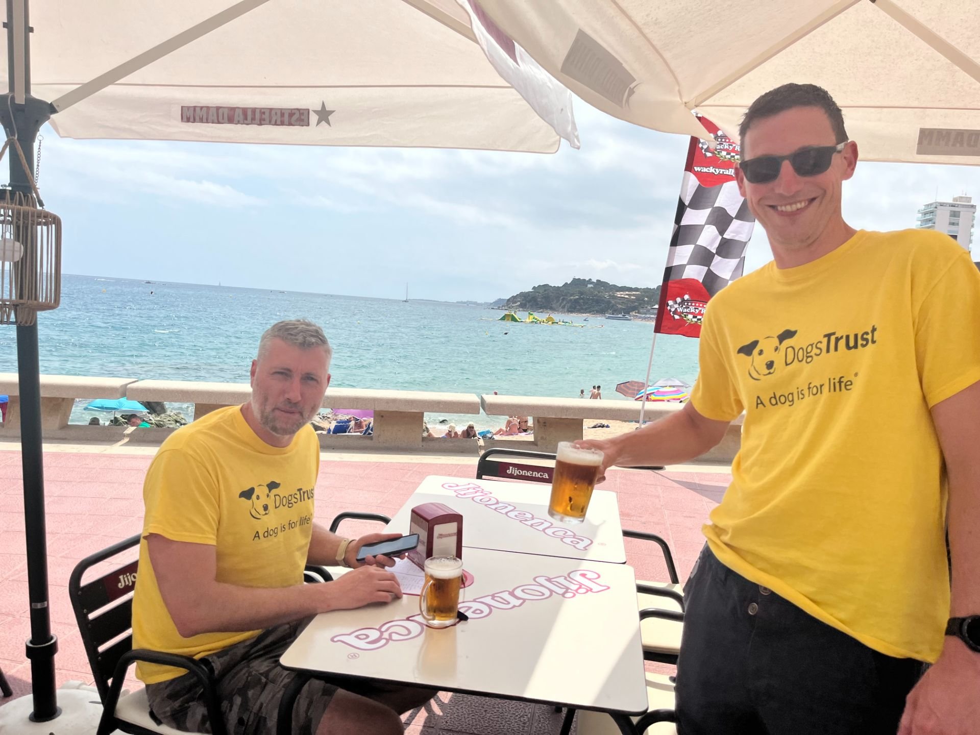 Two men in yellow t-shirts with DogsTrust logos sitting at an outdoor beachside cafe table, one holding a glass of beer, with a view of the beach and ocean in the background.