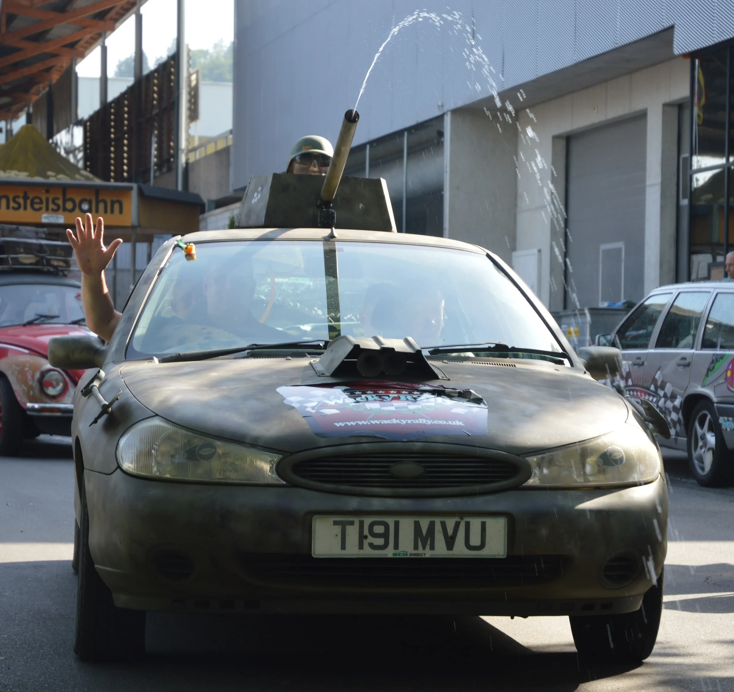 A modified, military-style vehicle with a mounted water cannon on top, spraying water. A person is waving out of the passenger window, and there are other cars behind it, including a pink car and a grey car with a checkered flag pattern. The vehicle 