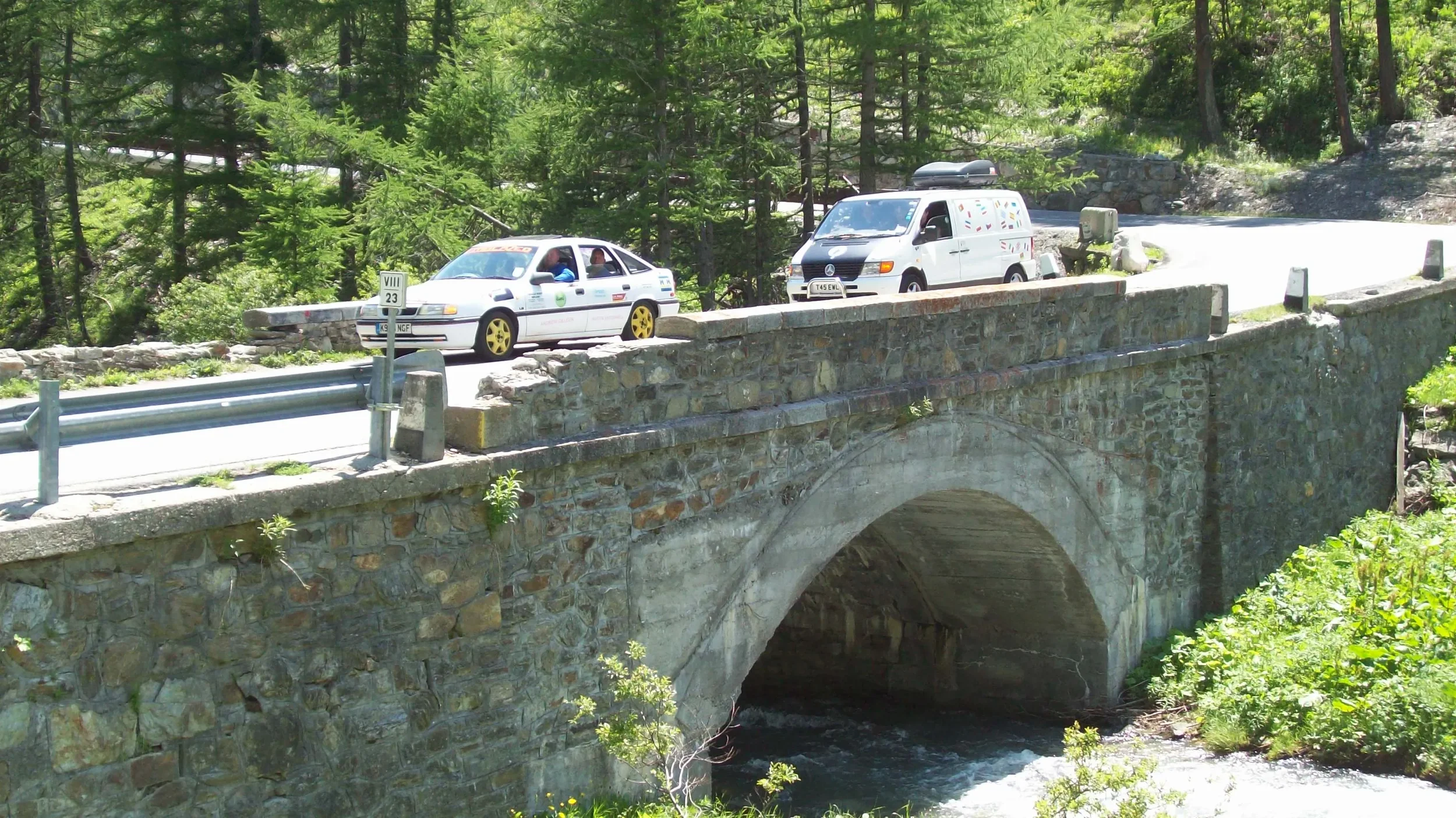 A stone bridge over a flowing stream surrounded by green trees and bushes, with a white car and a van parked on the bridge.
