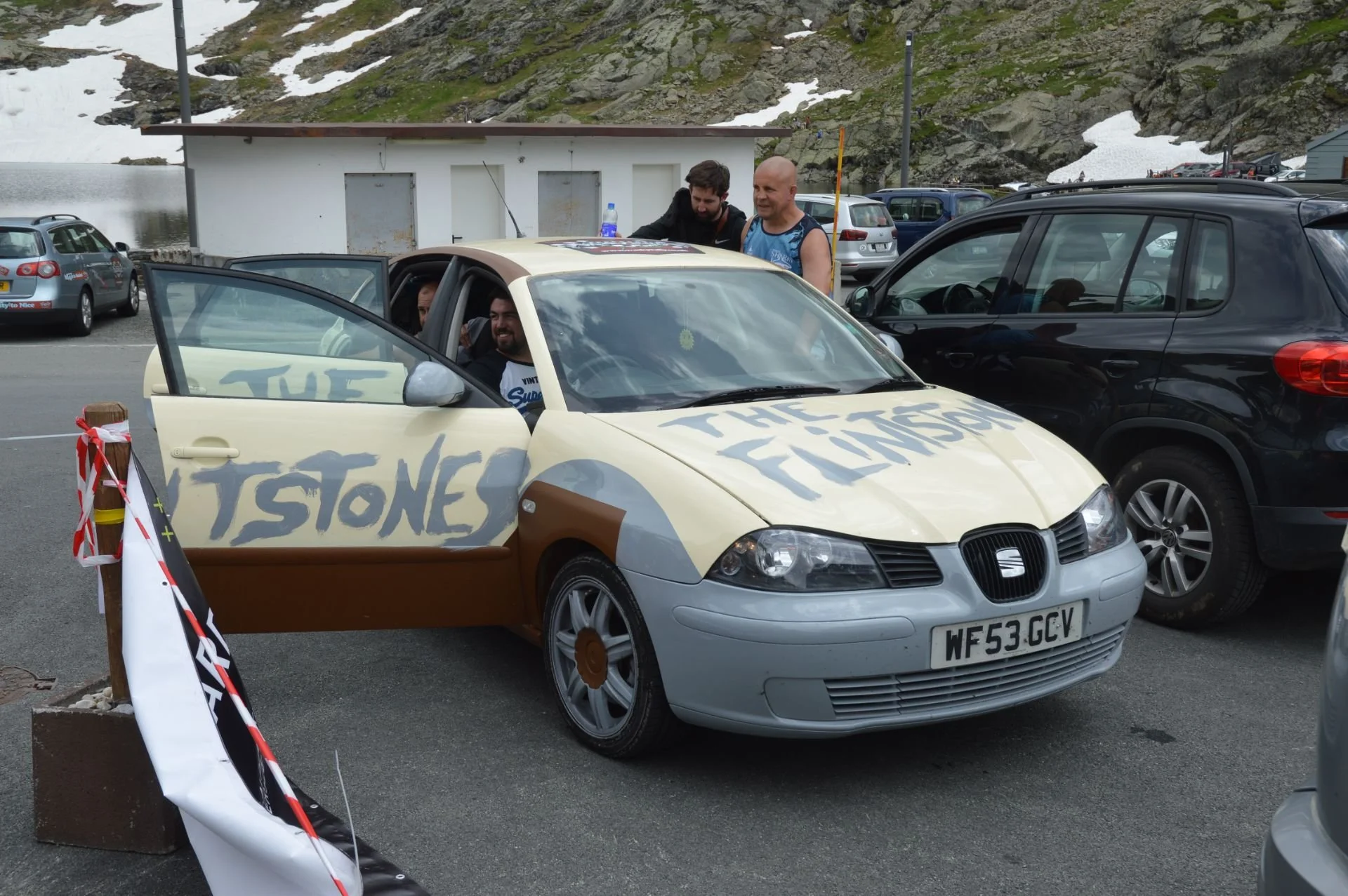 A decorated beige and gray Seat car with the words 'TSTONES' and 'THE FLATSONS' painted on it, parked in a lot with four men around it, one sitting inside. The parking lot is near a mountain with patches of snow and a small building in the background