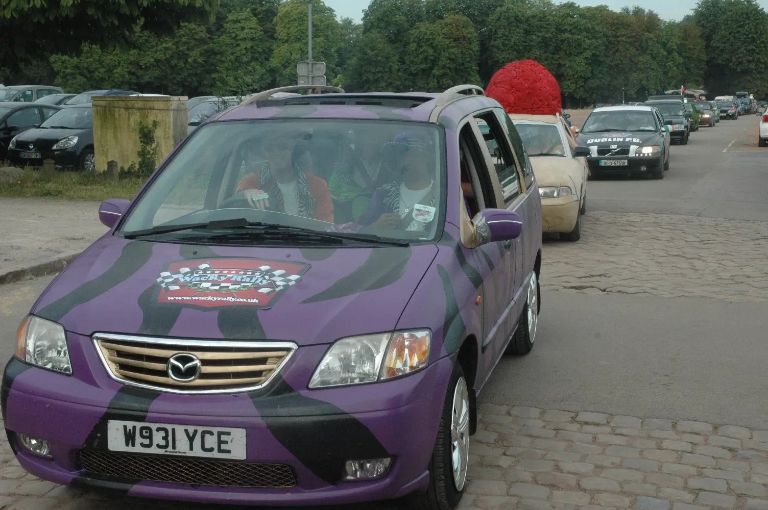 Purple Mazda car with a 'Wacky Rally' logo on the hood, parked on a cobblestone street, with people inside and line of parked cars in background.