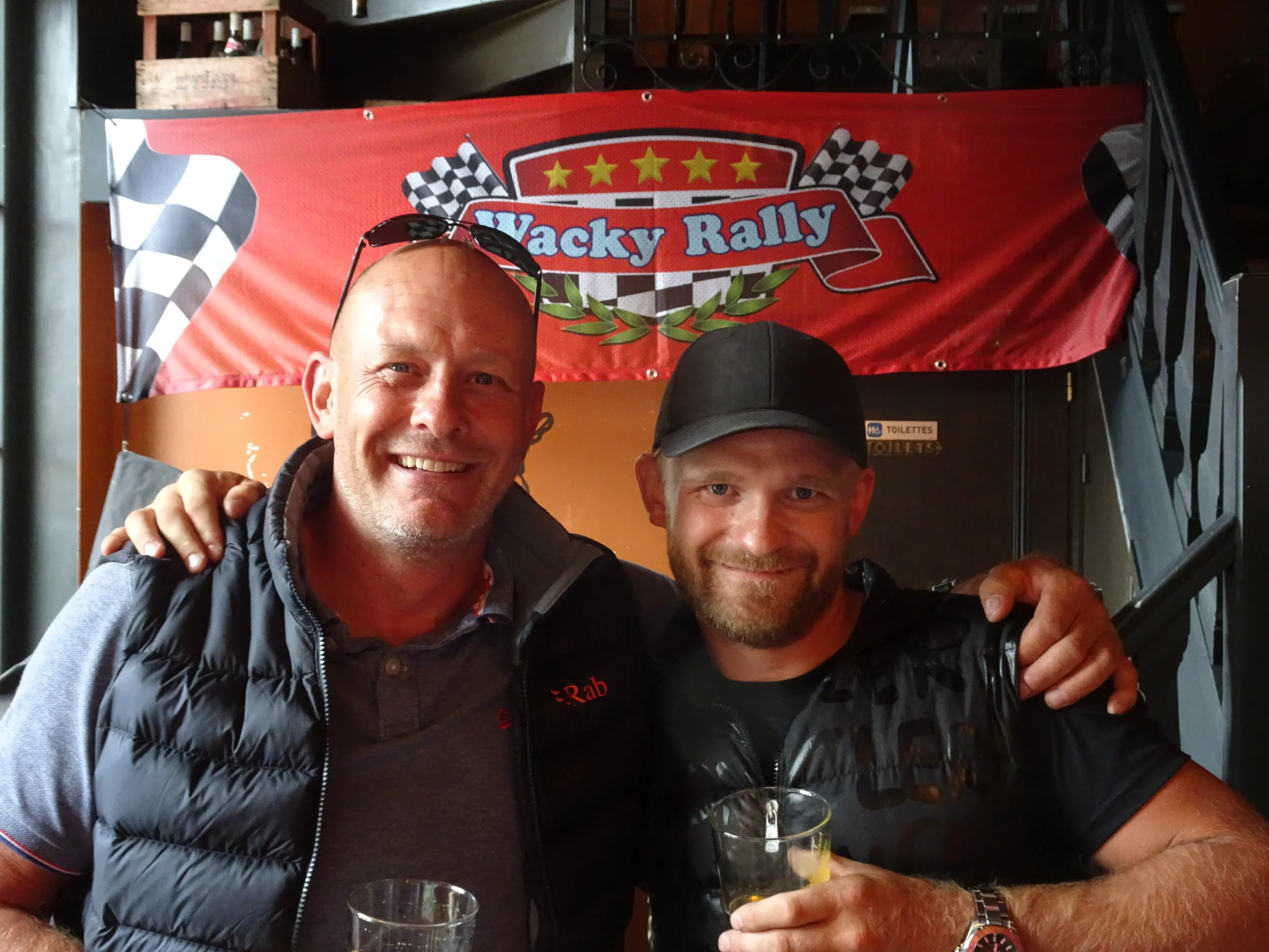 Two men smiling and posing together at a rally event with a red banner reading "Wacky Rally" and checkered flags in the background. One man has glasses on his head, and the other is wearing a black cap and holding a drink.