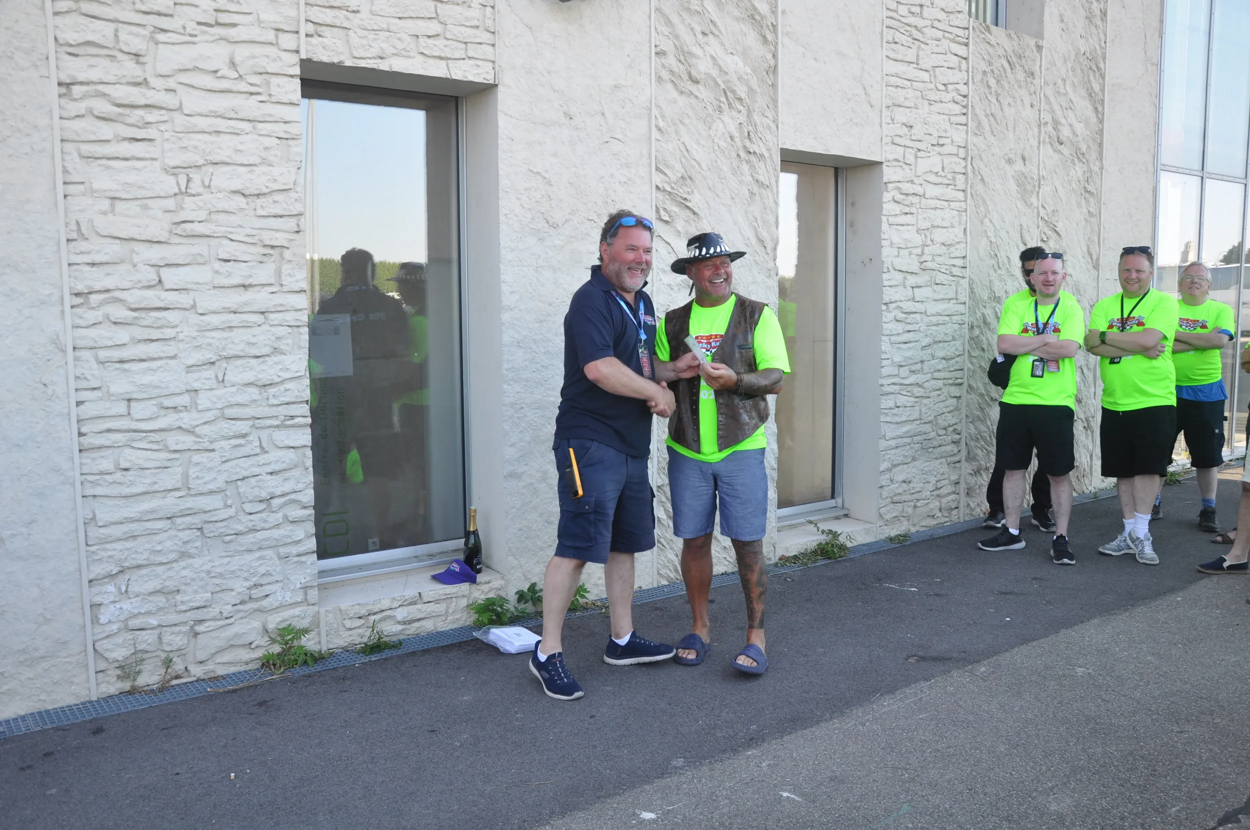 Two men standing in front of a stone building smiling and exchanging a card or paper, with three men in neon yellow shirts watching in the background.