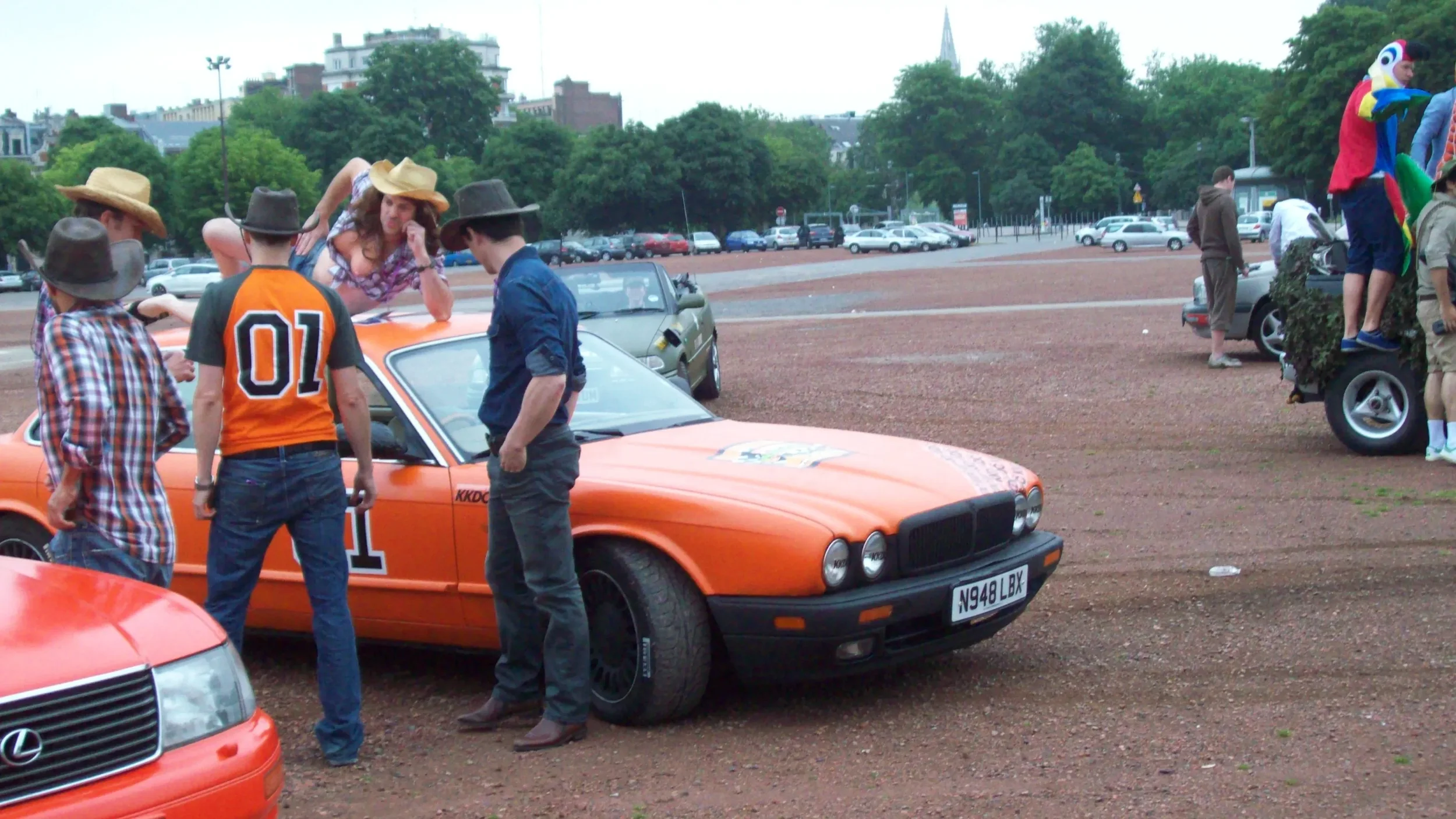 A group of people dressed in cowboy hats and casual clothing gathered around an orange vintage car with the number 01 on the side, parked on a gravel lot. A woman is leaning on the car, talking to the others, and there are other vintage cars and a fl