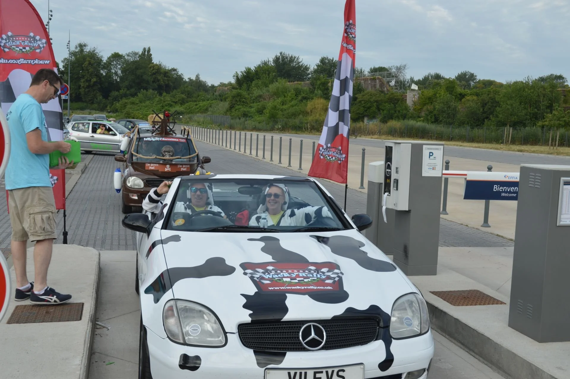 A white Mercedes convertible with a cow print design and a rally logo on the hood is in front of a parking ticket machine, with two smiling people inside wearing headphones. A man in a light blue t-shirt and khaki shorts stands nearby, holding a gree