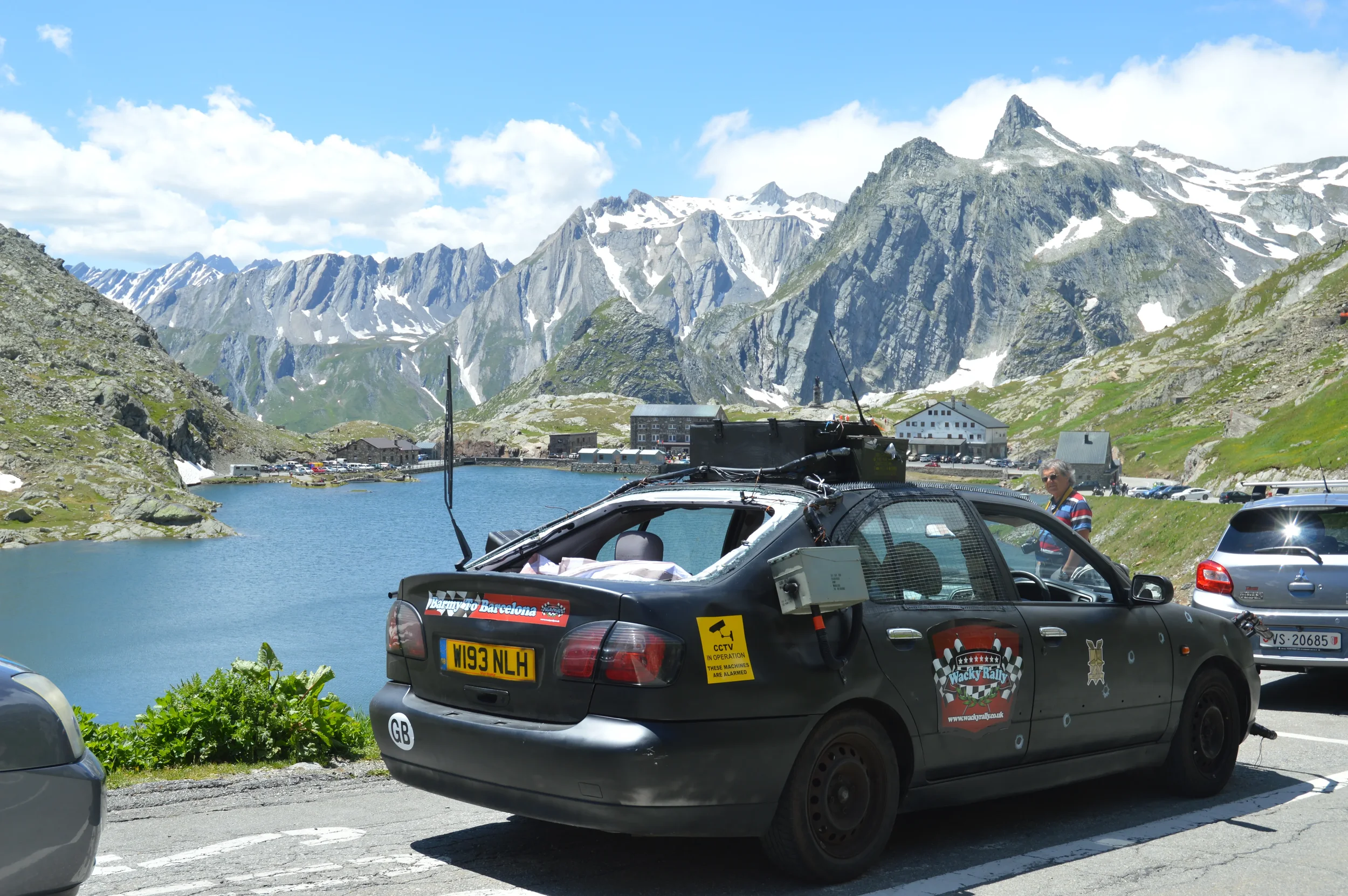 Black rally car parked by a mountain lake with snow-capped peaks in the background, surrounded by parked cars and a person standing nearby.
