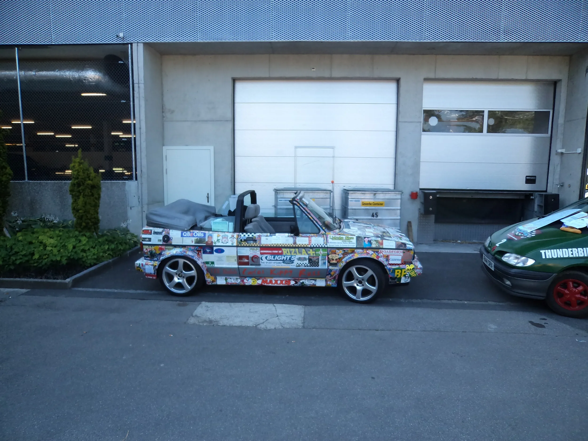 A small car covered with colorful stickers and decals, parked outside a building with white garage doors and a glass window, next to a green car with red wheels.