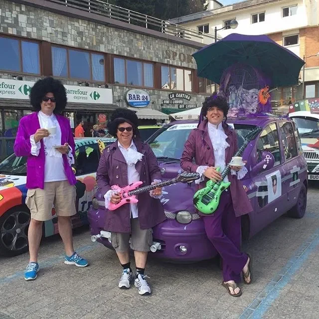 Three people dressed as The Beatles from Sgt. Pepper's Lonely Hearts Club Band stand in front of a purple car decorated with musical instruments, umbrellas, and purple ribbons, on a city street during a parade or event.