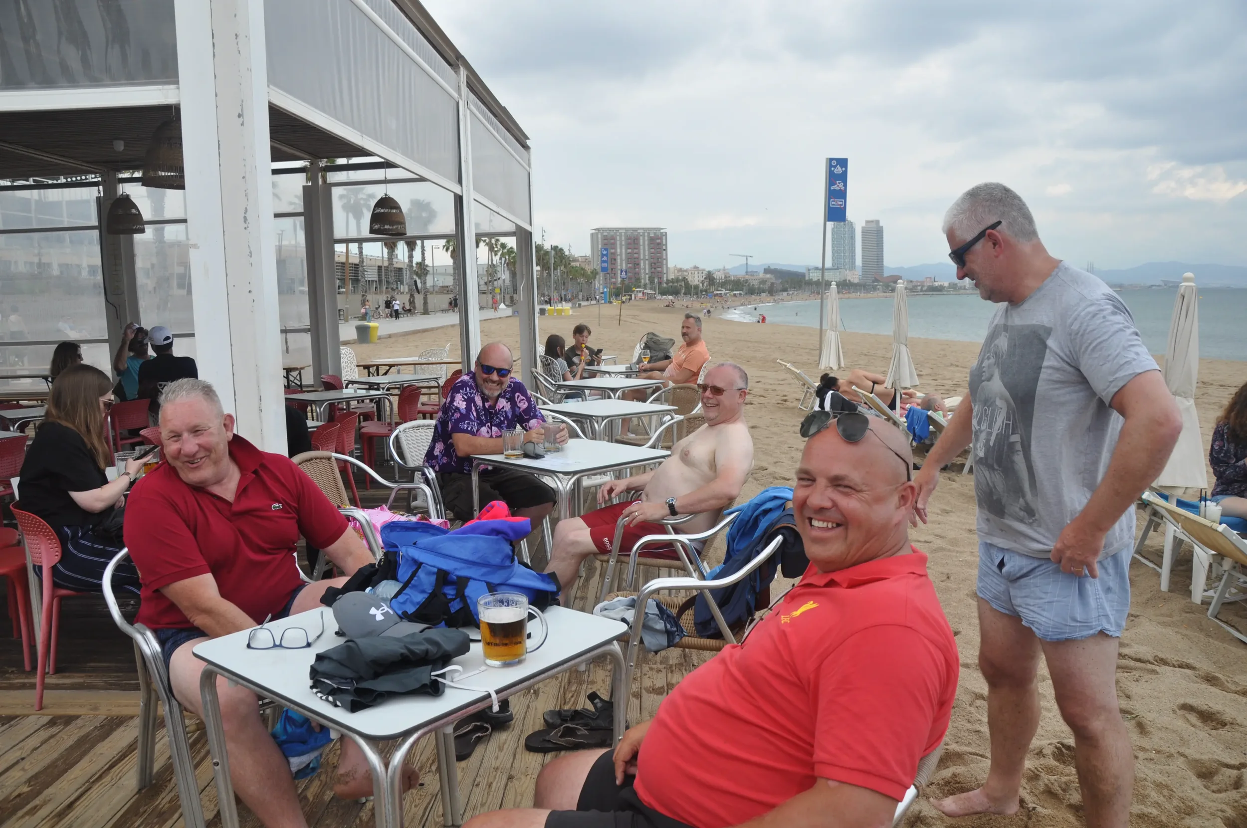 Group of friends at an outdoor beachside restaurant, smiling and socializing, with beach and city skyline in the background.