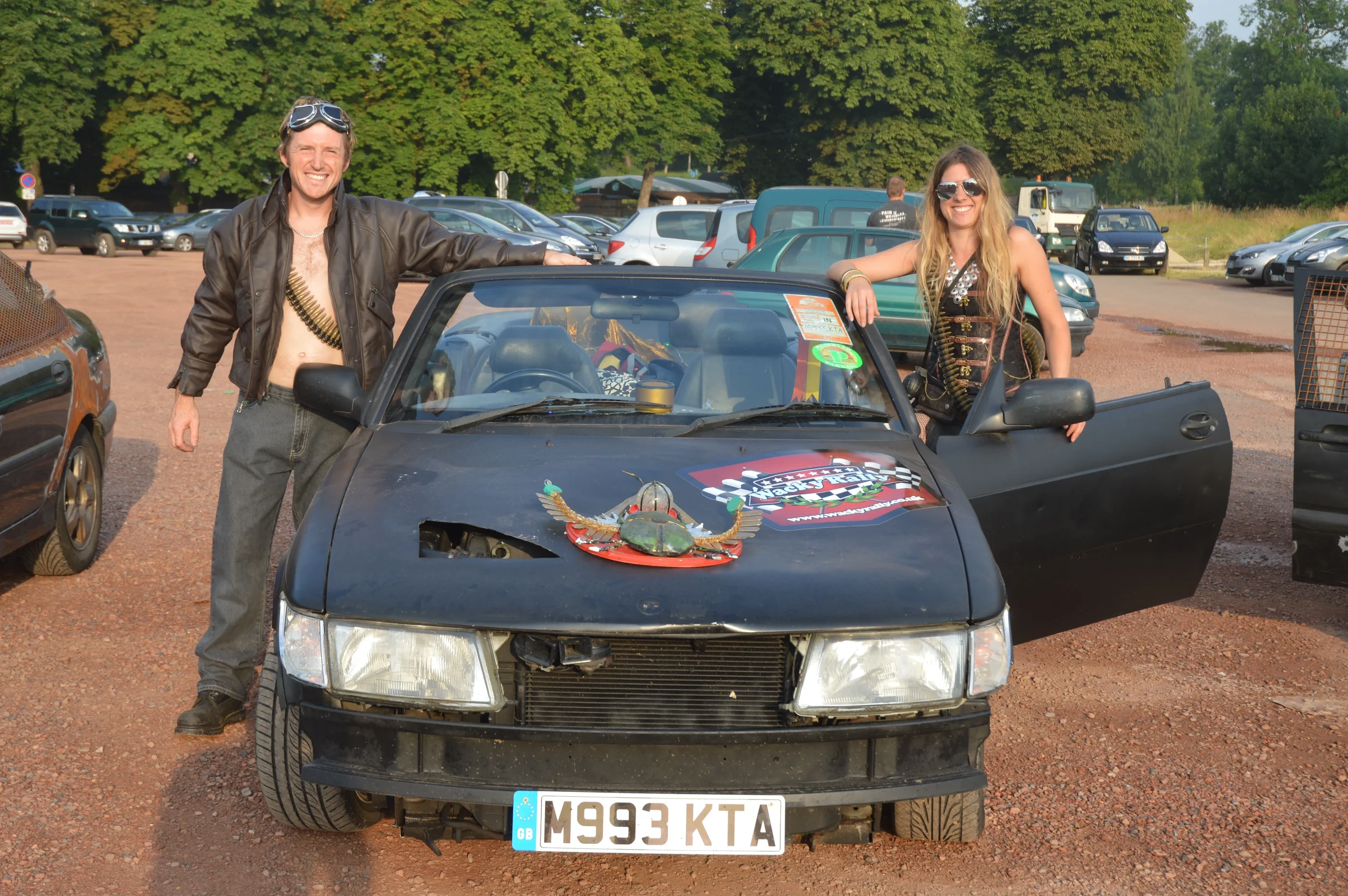 Two smiling people, a man and a woman, posing next to a black car in a parking lot. The man is shirtless with a leather jacket and goggles, and the woman is wearing sunglasses and a sleeveless top. There are cars and trees in the background.