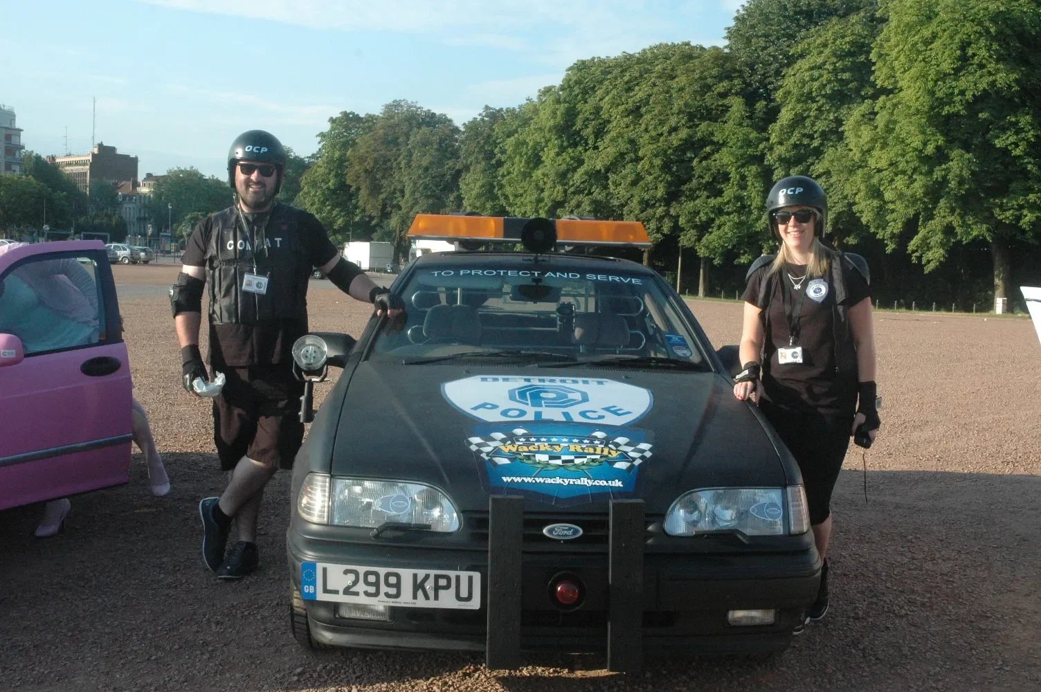 Two police officers in black uniforms and helmets standing beside a black police car marked with logos and a website URL. The officers are smiling and posing for the camera. The background shows trees, a gravel area, and some buildings in the distanc