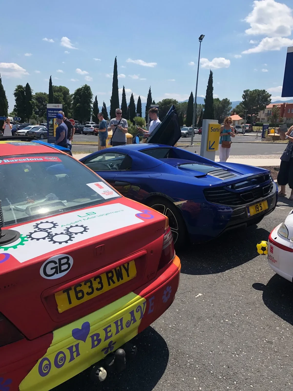Several cars parked in an outdoor lot with people standing around. One car is blue with its engine cover open, and another is red with motivational stickers on the back. The scene appears to be at a car show or gathering on a sunny day with trees and