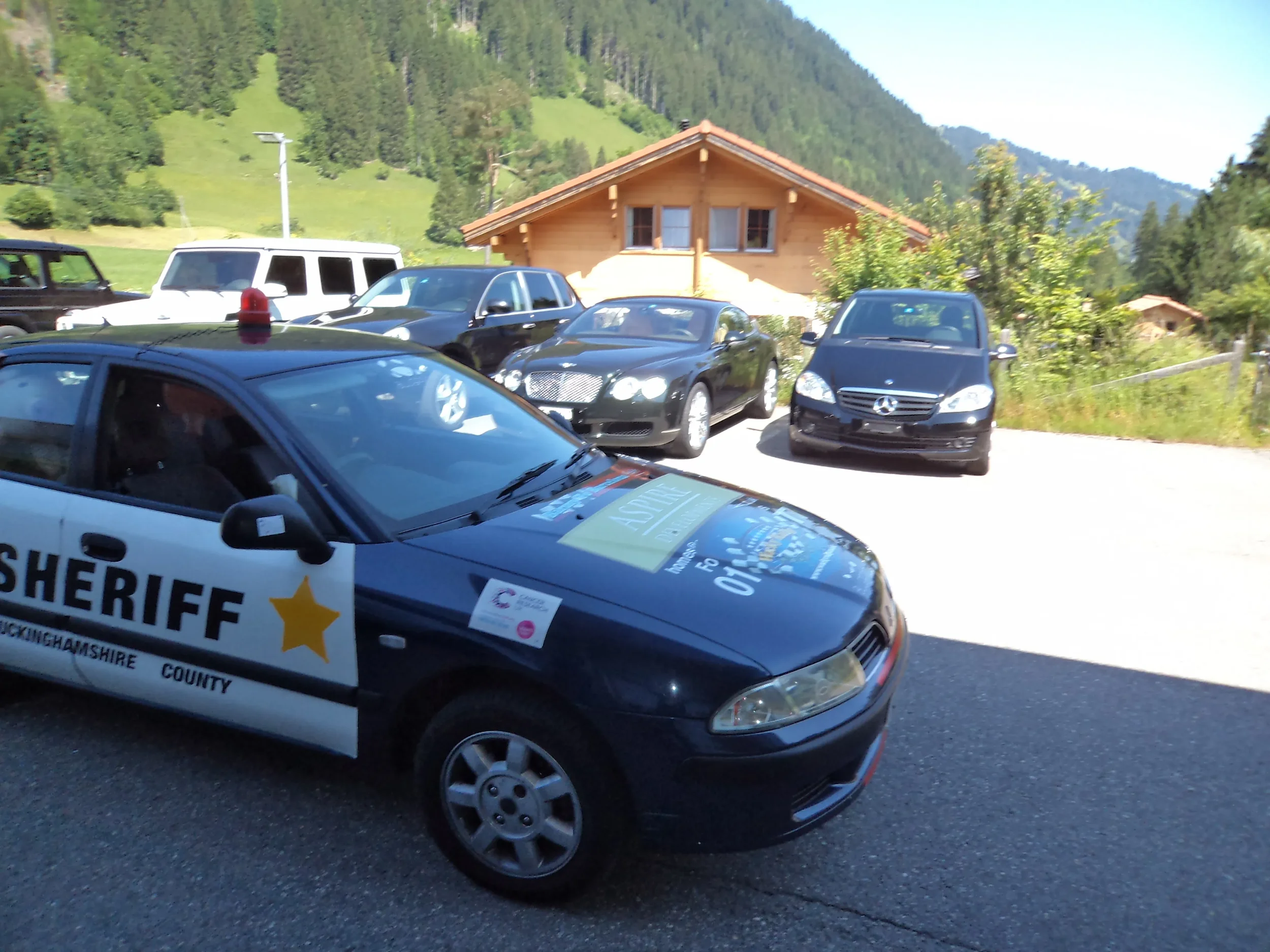 A police sheriff car parked in front of a house in a mountainous area with several other parked cars around, including a black Mercedes-Benz and a white vehicle, with green trees and hills in the background.