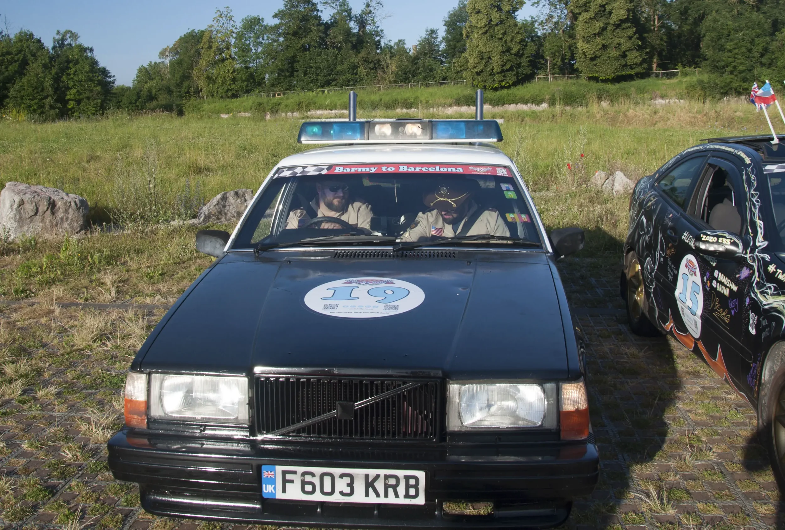 Black vintage Volvo car with racing number 119 on the hood, parked on a grassy area with rocks and trees in the background. Two men dressed as vintage race drivers sit inside. A banner on the windshield reads 'Barmy to Barcelona,' and the car has a B