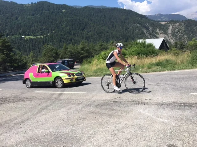 A cyclist riding a bike on a road with a colorful car in the background and mountains in the distance.