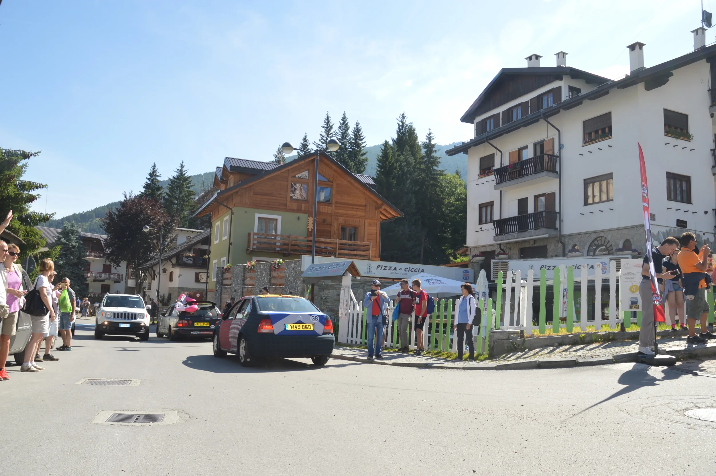 Tourists standing and walking on a street in a mountain village with colorful buildings and parked cars, sunny day.