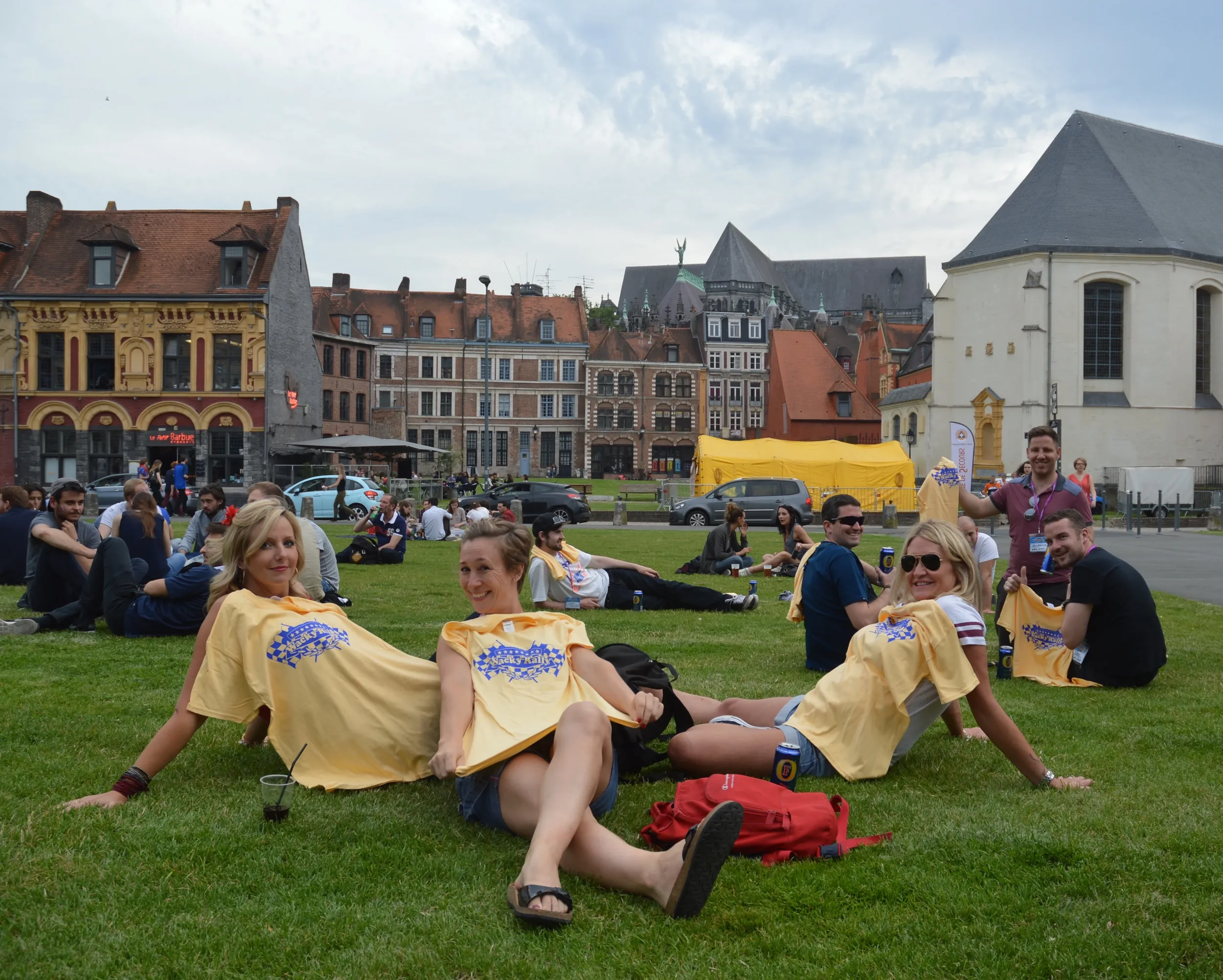 Group of friends sitting and relaxing on a grassy field, some wearing matching yellow shirts, with historical buildings and a cloudy sky in the background.