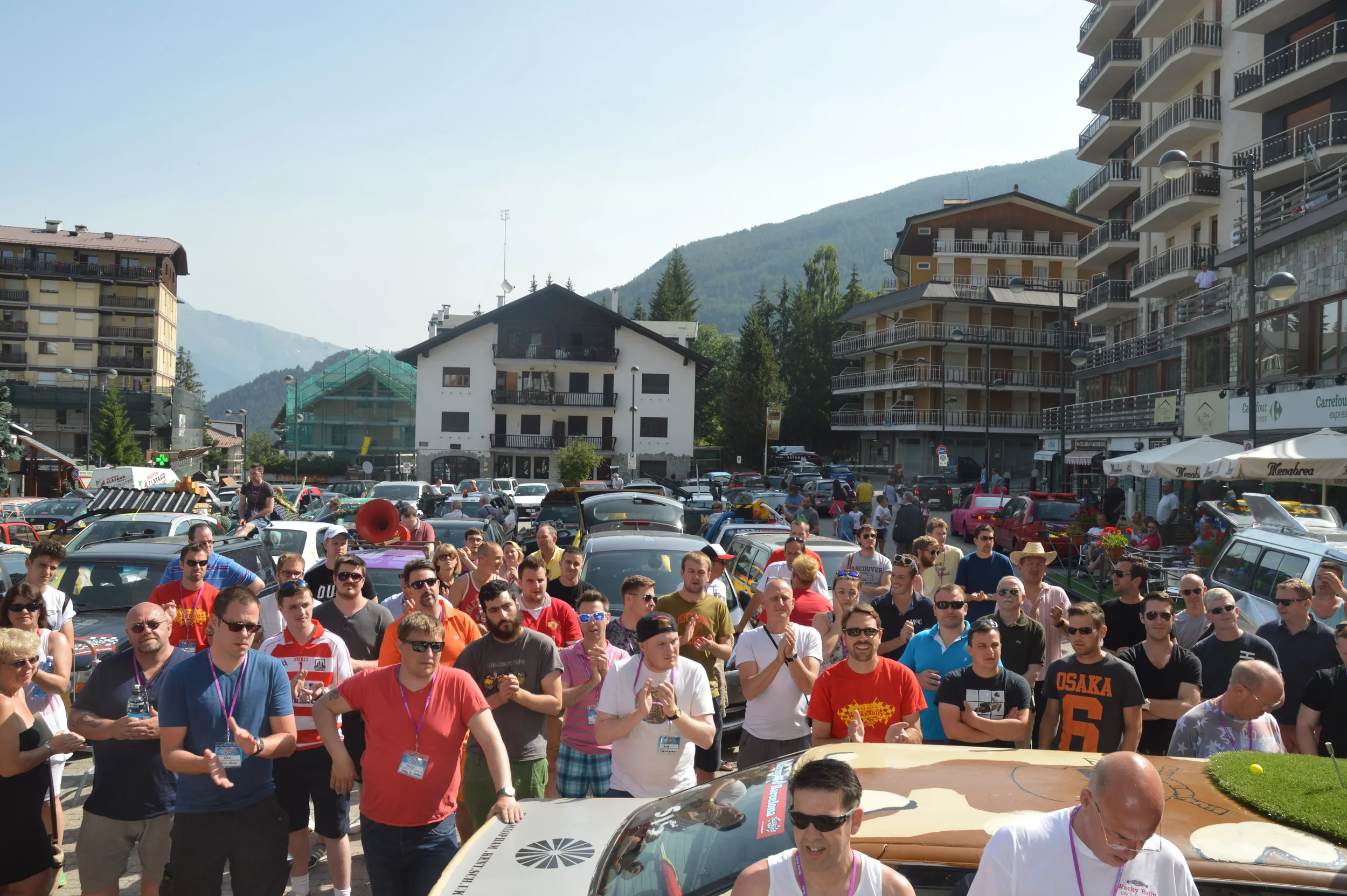 Crowd of people gathered outdoors with mountains in the background, cars parked nearby, some onlookers on balconies of buildings, sunny weather.