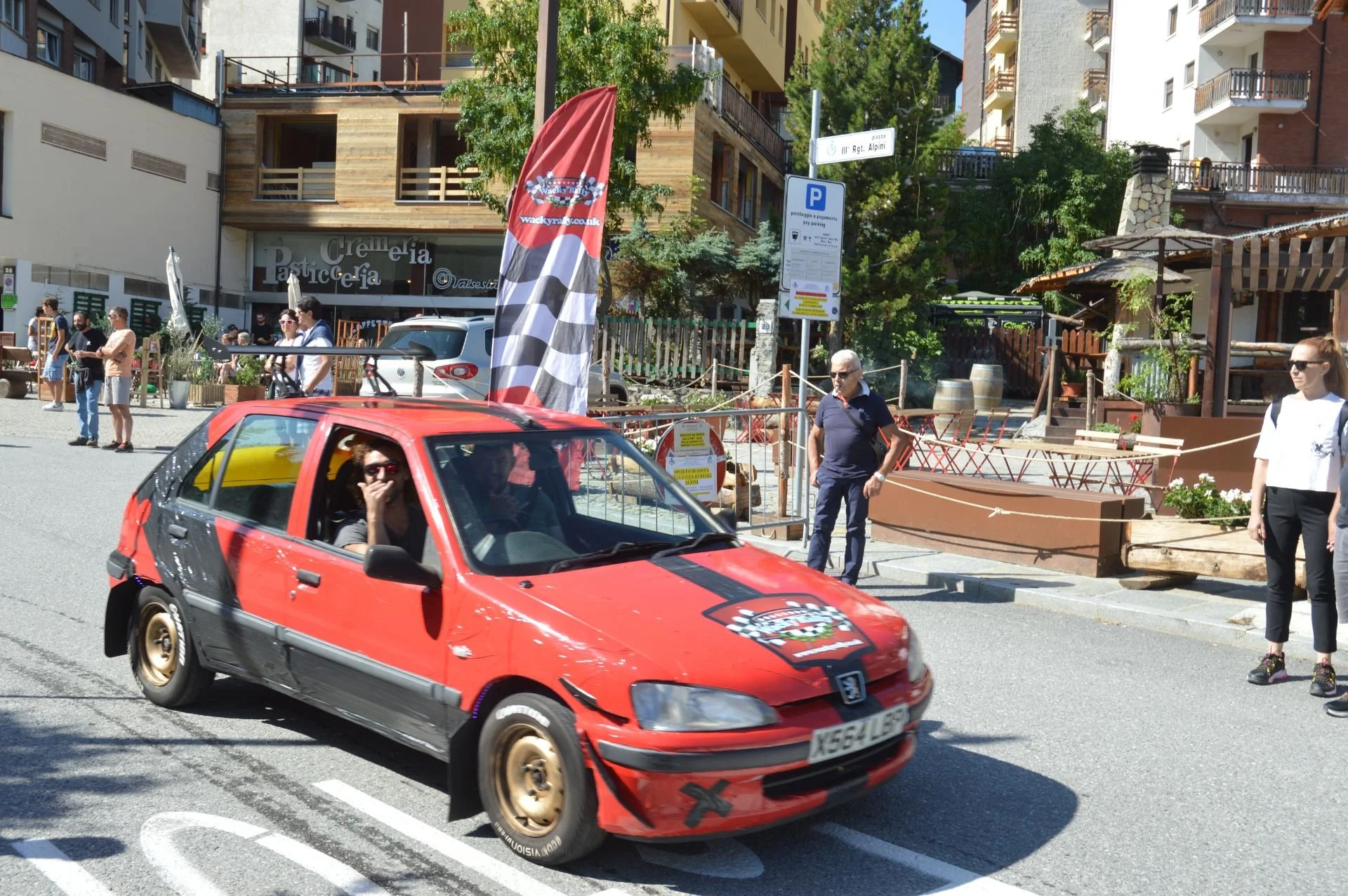 Red Peugeot rally car with a checkered flag decal, driving through a street with spectators and buildings in the background.