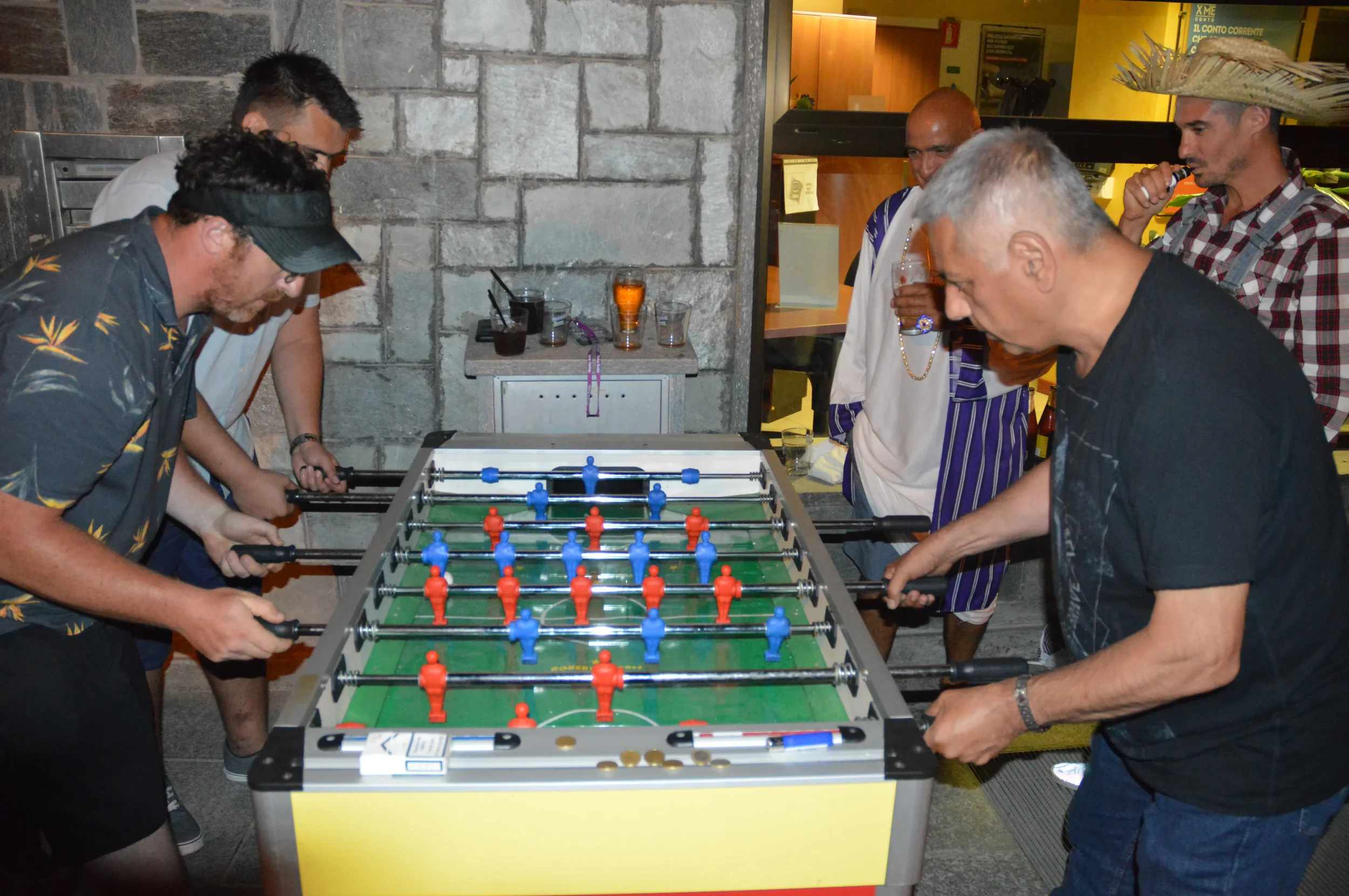 Group of people playing foosball in a room with stone wall and drinks on a table.