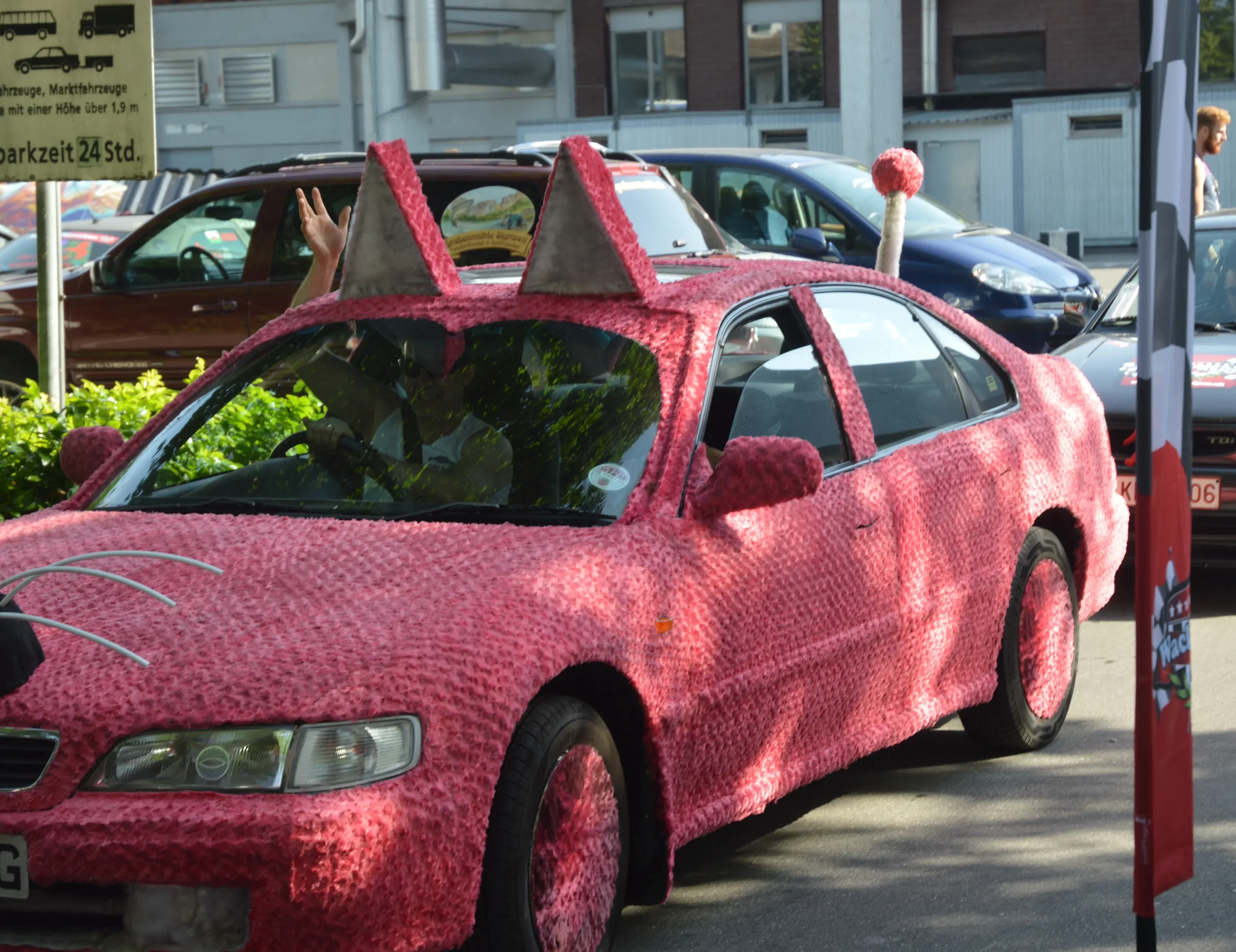 Pink car decorated with fuzzy, textured material resembling craft or costume fabric, featuring cat ears on the roof and a pink pom-pom tail on the trunk, parked in an outdoor lot.