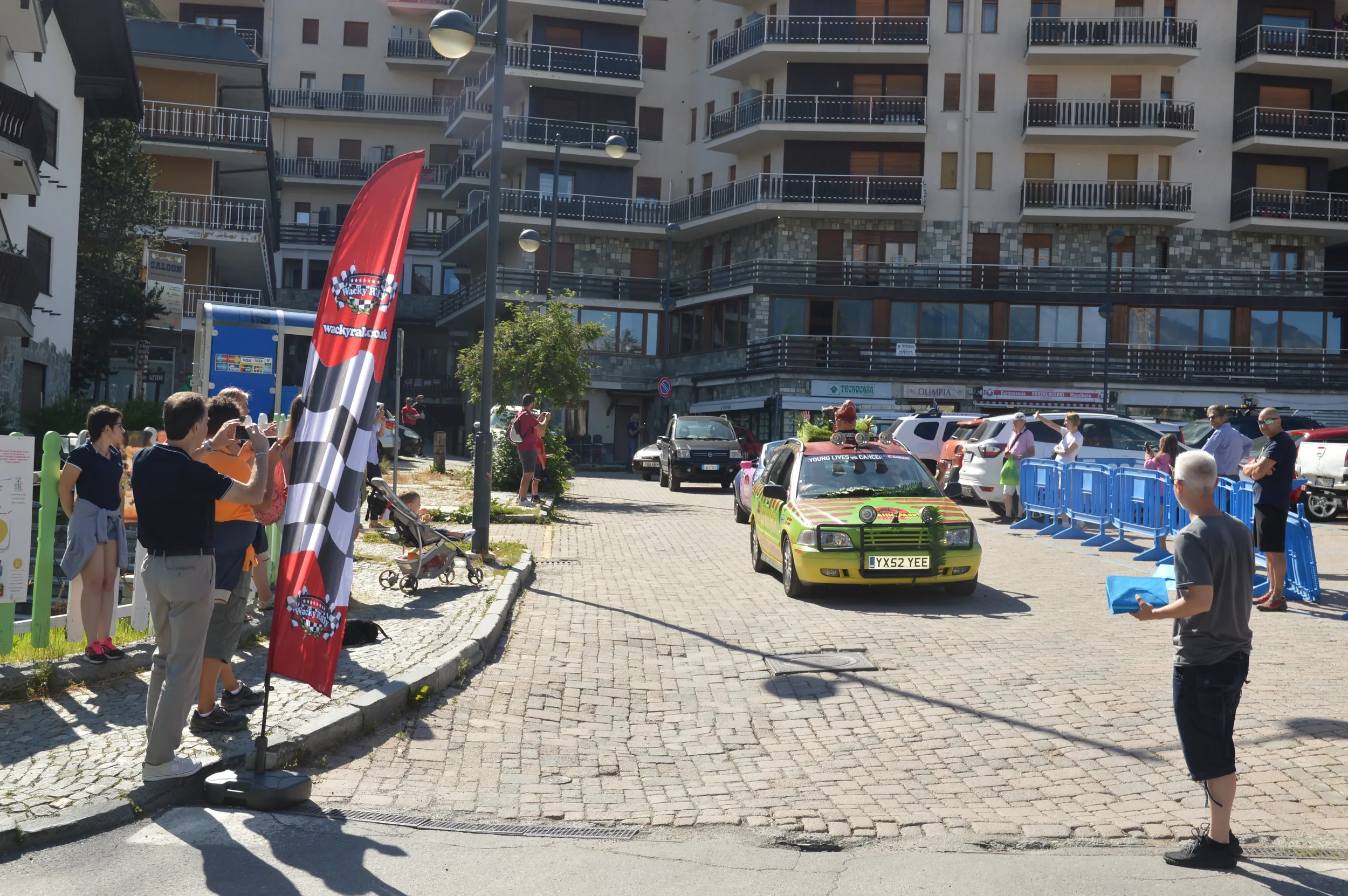 A small parade of colorful cars driving on cobblestone street, lined with spectators taking photos, young and old, surrounded by modern apartments with balconies and parked cars.
