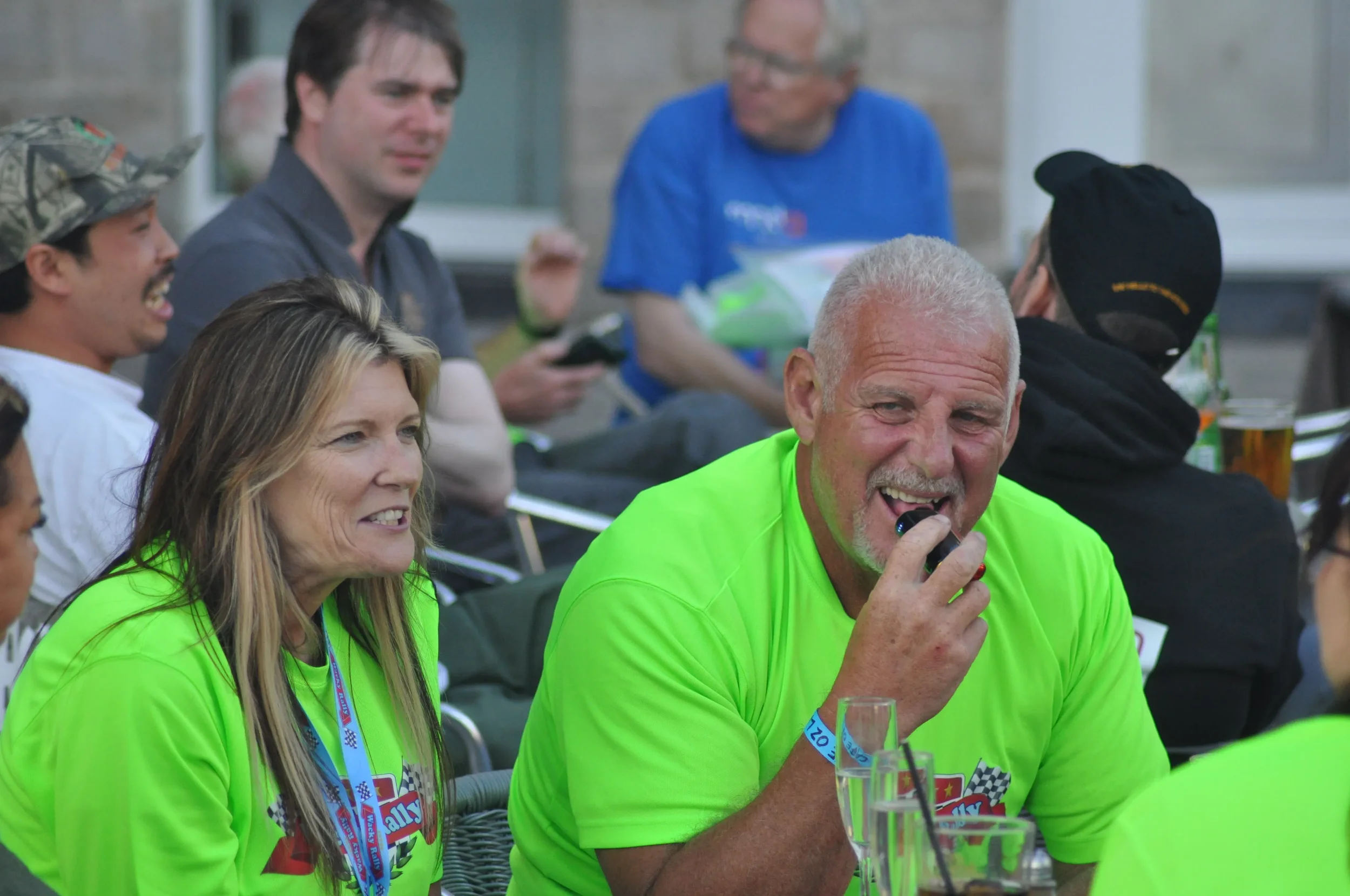 A group of people sitting at an outdoor gathering, with a man in a bright green shirt smiling and holding a mouthpiece, and a woman in a matching shirt smiling beside him, surrounded by others in casual attire.